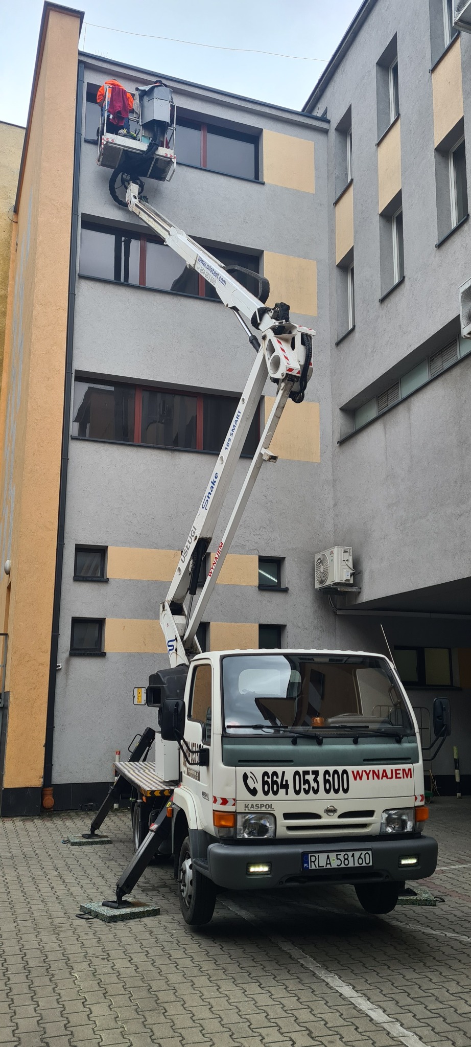 Podnośnik koszowy na tle budynku. Dwóch pracowników w pomarańczowych ubraniach w koszu podnośnika. Na pojeździe widoczny numer telefonu i napis 'WYNAJEM'.