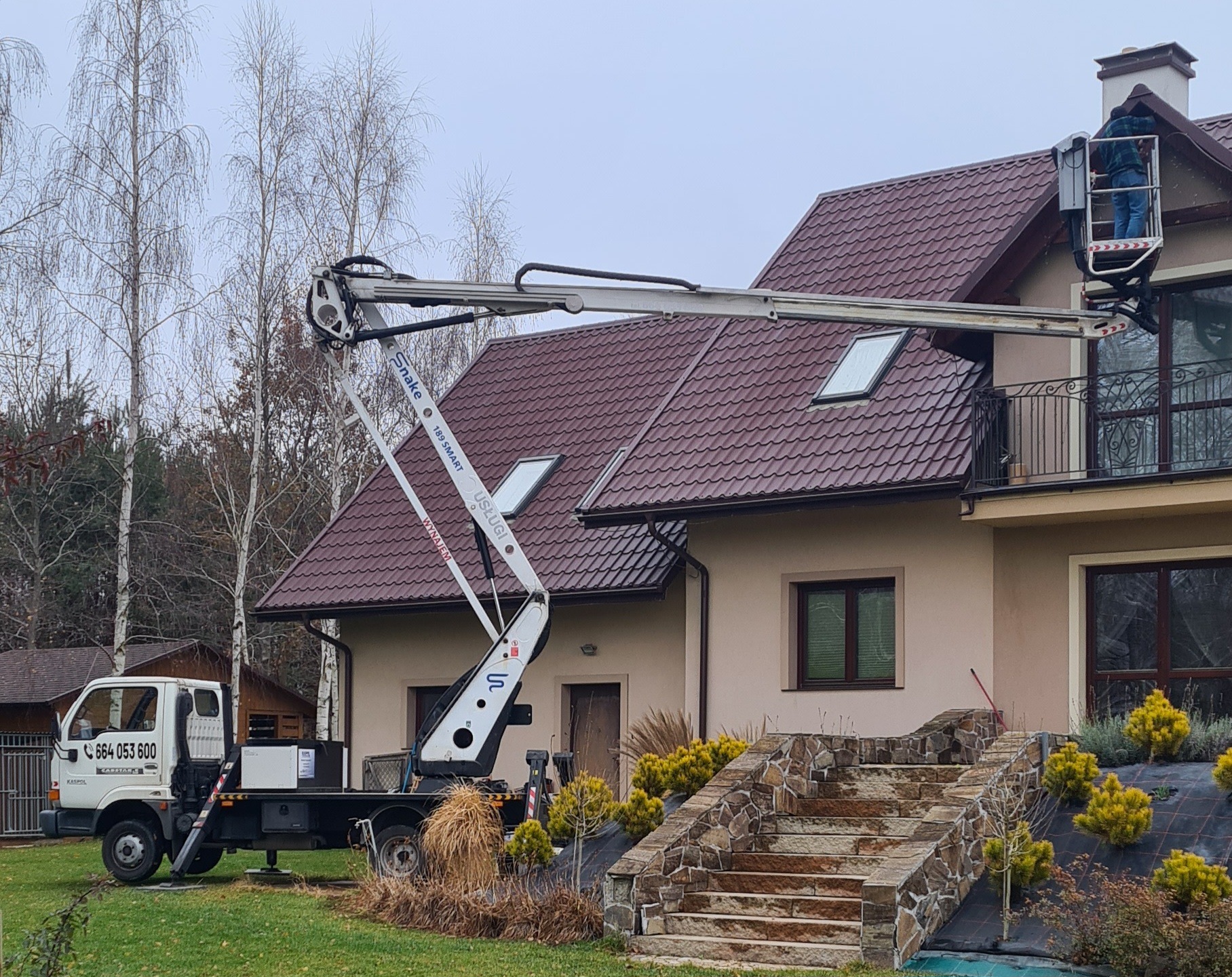 Pracownik w koszu podnośnika koszowego przy dachu domu z brązową dachówką. Podnośnik marki Snake, biała elewacja. Widoczne schody wejściowe z kamienia.