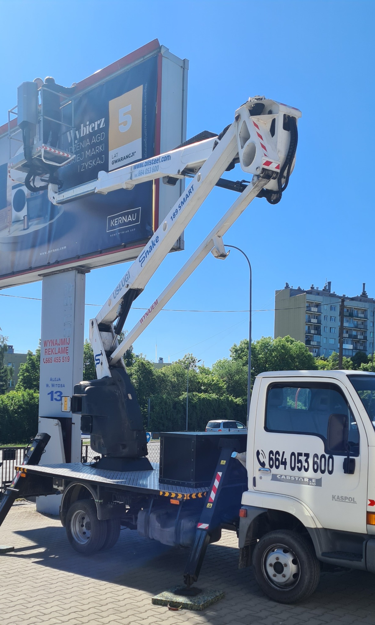 Podnośnik koszowy Snake 189 Smart na tle błękitnego nieba, pracujący przy montażu baneru reklamowego na wysokości. Widoczne numery kontaktowe na pojeździe.