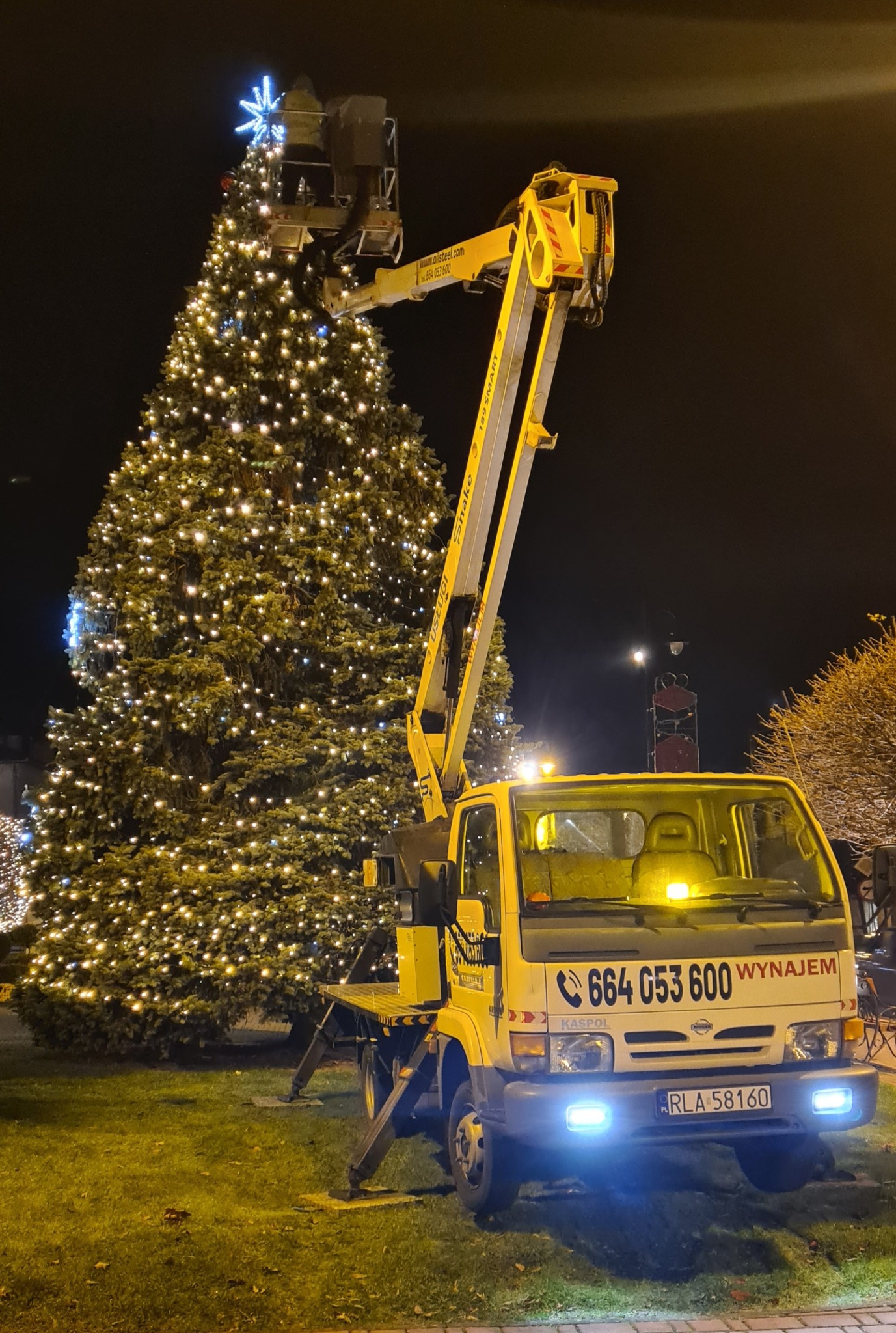 Żółty podnośnik koszowy na tle oświetlonej choinki miejskiej z gwiazdą na szczycie, nocą. Widoczny numer kontaktowy na pojeździe.