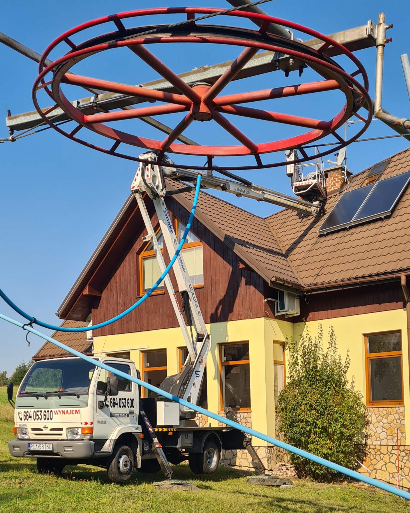 Podnośnik koszowy na tle domu z panelami słonecznymi na dachu. Widoczny czerwony element konstrukcyjny nad wysięgnikiem. Żółta elewacja budynku.