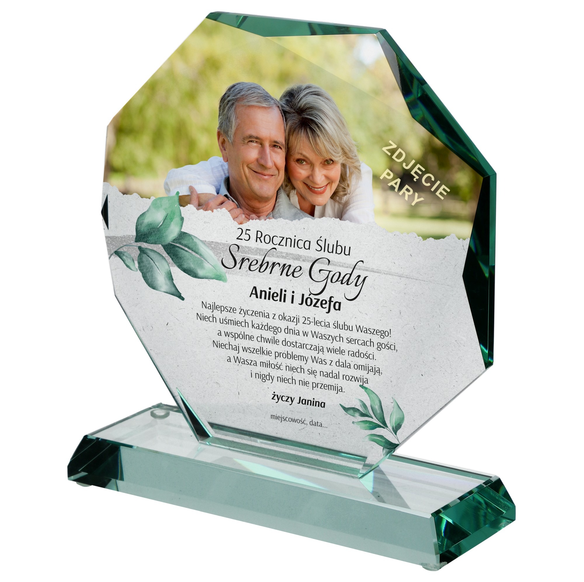 Szklana statuetka z nadrukiem zdjęcia pary i życzeń z okazji 25. rocznicy ślubu, umieszczona na przezroczystej podstawie, z widoczną zielenią w tle.