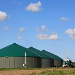 Kompleks biogazowni z zielonymi kopułami na tle błękitnego nieba z chmurami, otoczony polami i trawą. Widoczne zbiorniki i instalacje technologiczne.