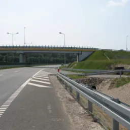 Autostrada z wiaduktem nad drogą, bariery ochronne i przepust dla zwierząt. Widoczna infrastruktura drogowa i fragment zieleni.