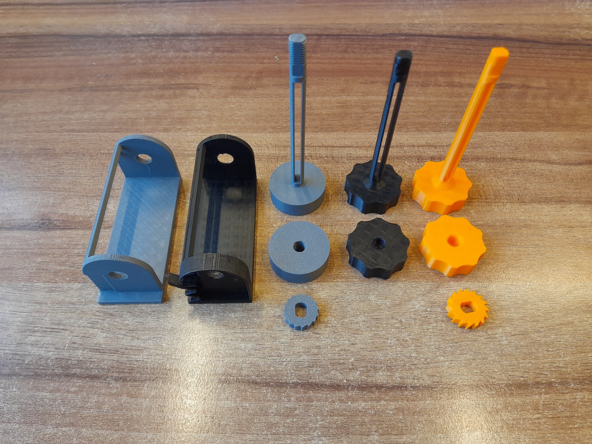 Różnokolorowe, plastikowe elementy, prawdopodobnie wydrukowane w 3D, ułożone na drewnianej powierzchni. Widoczne podpory, nakrętki i inne detale.