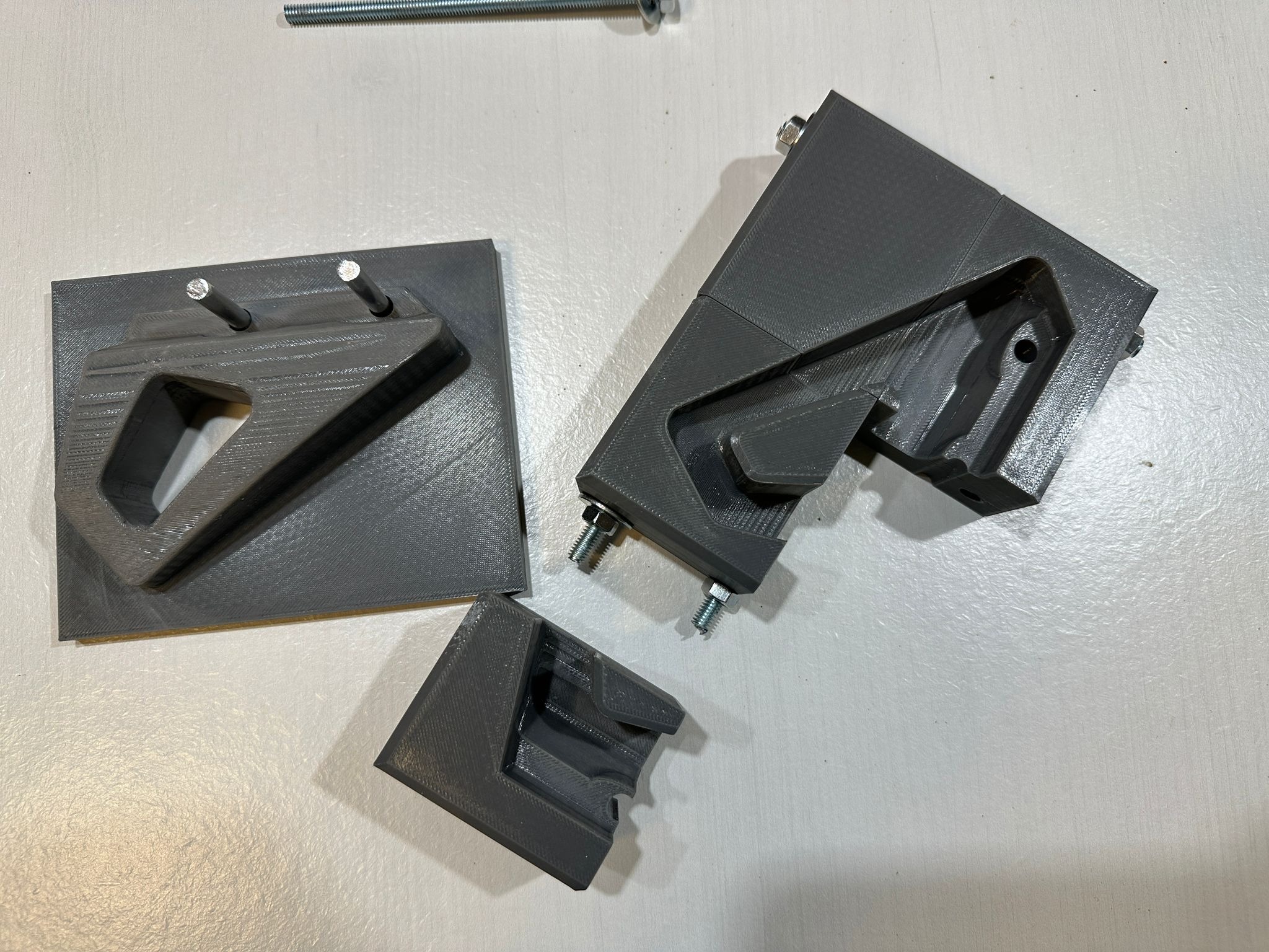 Szare, rozłożone elementy z tworzywa sztucznego, prawdopodobnie wydrukowane, z widocznymi śrubami i gwintowanym prętem, na jasnym tle.