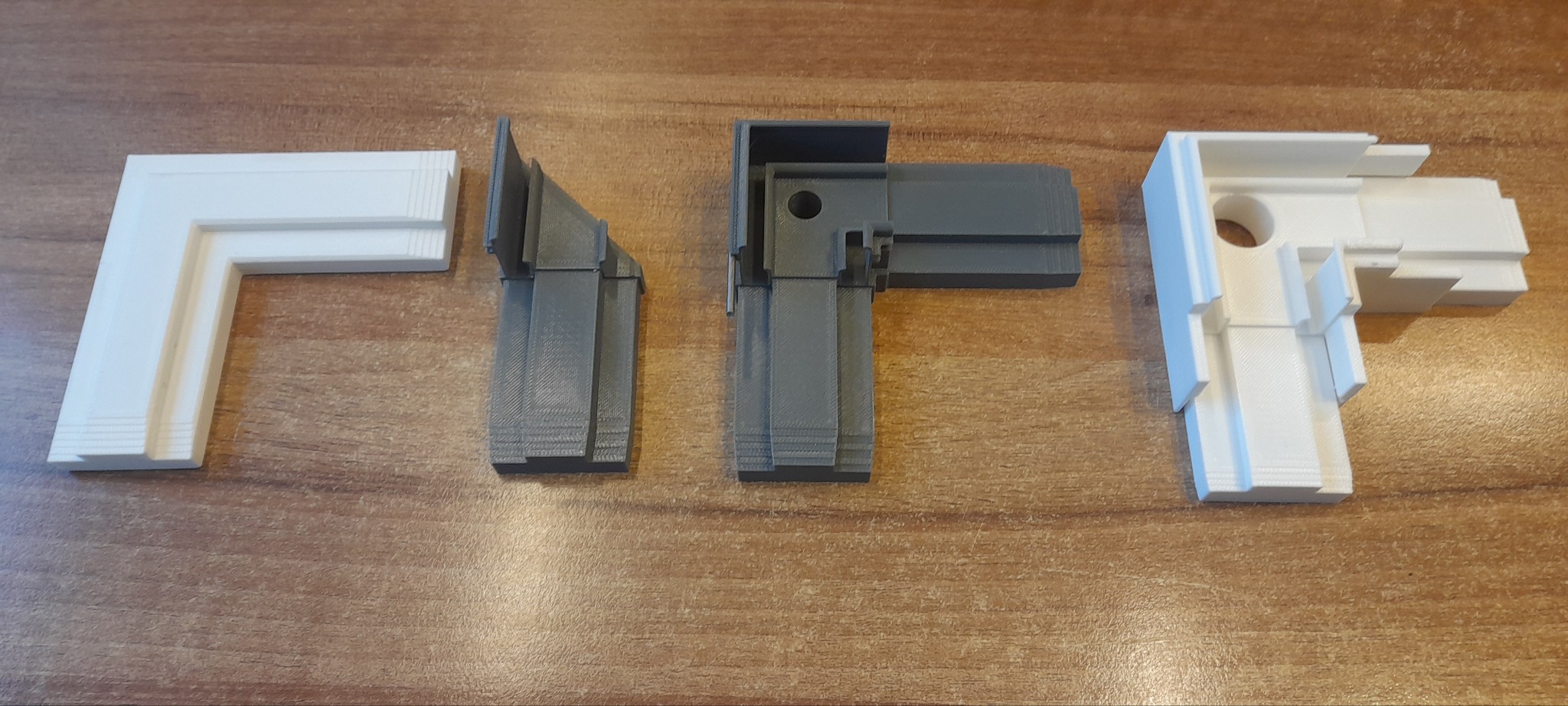 Cztery narożne elementy, wydrukowane w 3D w odcieniach bieli i szarości, leżą na drewnianej powierzchni. Widoczne warstwy druku.