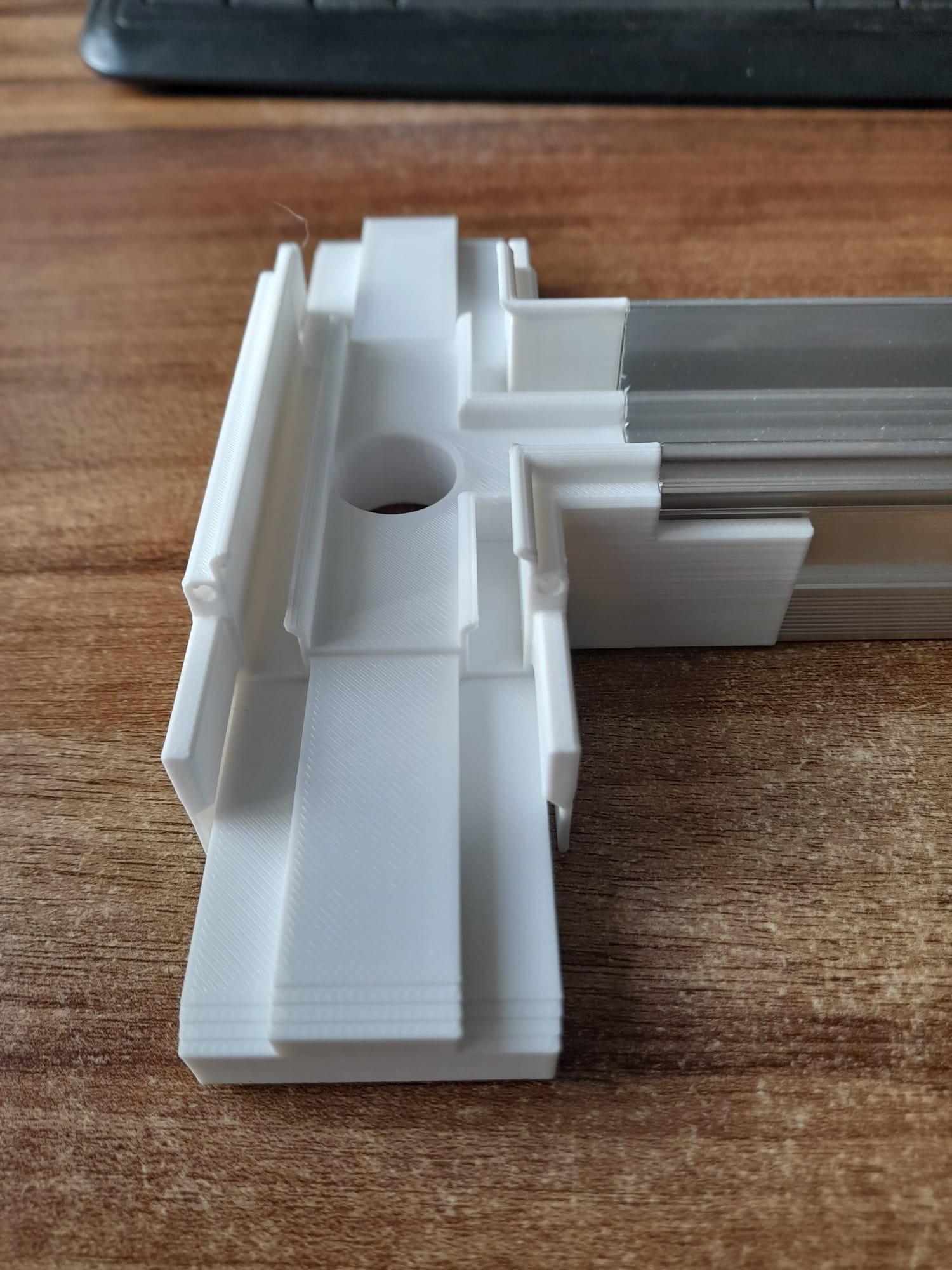 Biały, narożny element konstrukcyjny z widoczną strukturą druku 3D i metalową szyną, leżący na drewnianym blacie. Precyzyjne wykonanie detali z tworzywa sztucznego.