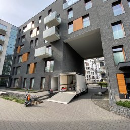 Jack Strong - Przeprowadzka w nowoczesnym apartamentowcu z szarej cegły. Biała furgonetka z rampą i paleciak elektryczny na brukowanym podjeździe.