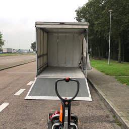 Jack Strong - Otwarta paka busa z rampą załadowczą i elektrycznym wózkiem paletowym na asfalcie, w tle zieleń i droga. Sprzęt gotowy do załadunku towarów.