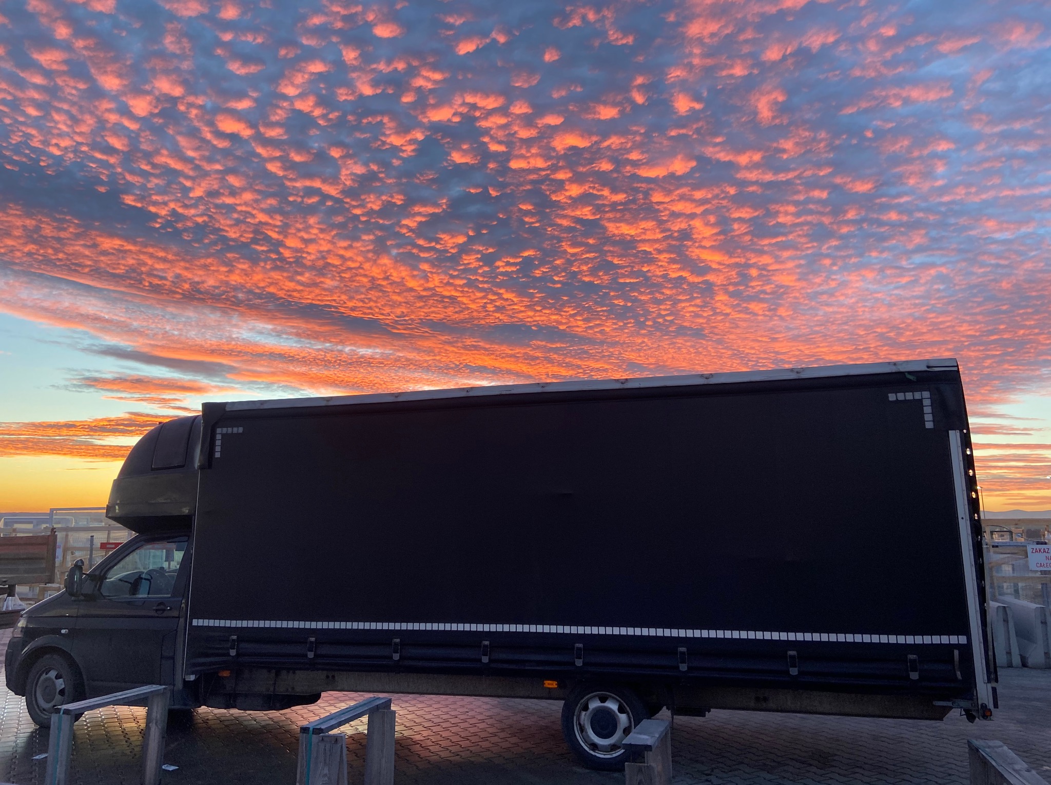 Czarny bus dostawczy na tle dramatycznego, pomarańczowo-różowego nieba o wschodzie słońca. Widoczne detale pojazdu i otoczenia.