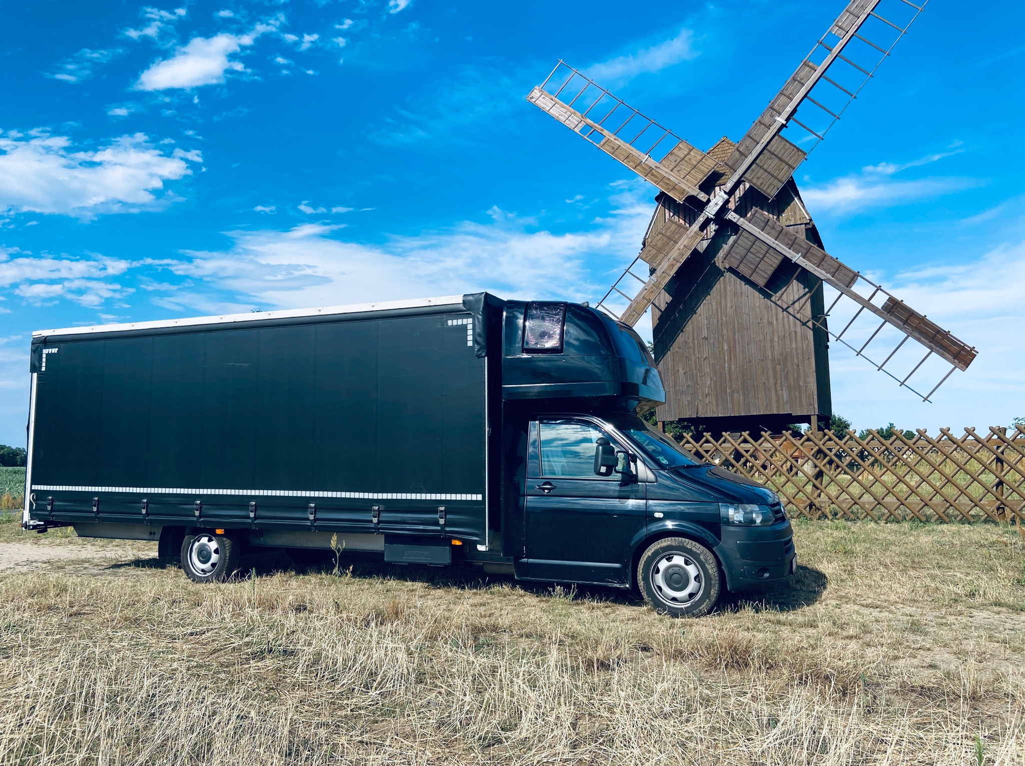 Czarny bus transportowy na tle wiatraka i błękitnego nieba z chmurami. Nietypowa lokalizacja prezentuje możliwości transportowe w różnych warunkach.