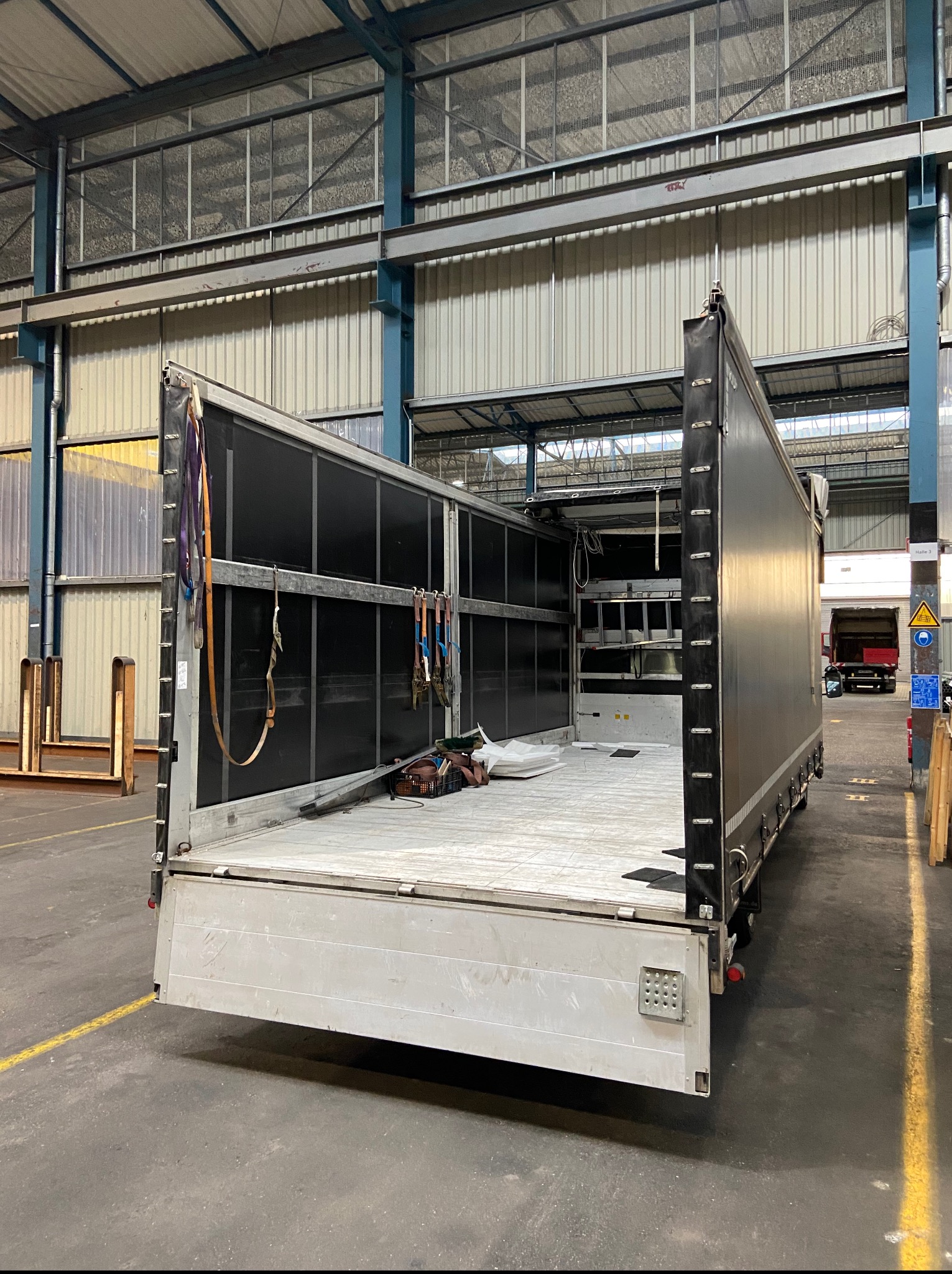 Półotwarta naczepa ciężarówki z widocznymi pasami mocującymi ładunek i podłogą, w hali magazynowej z żółtymi liniami na posadzce.