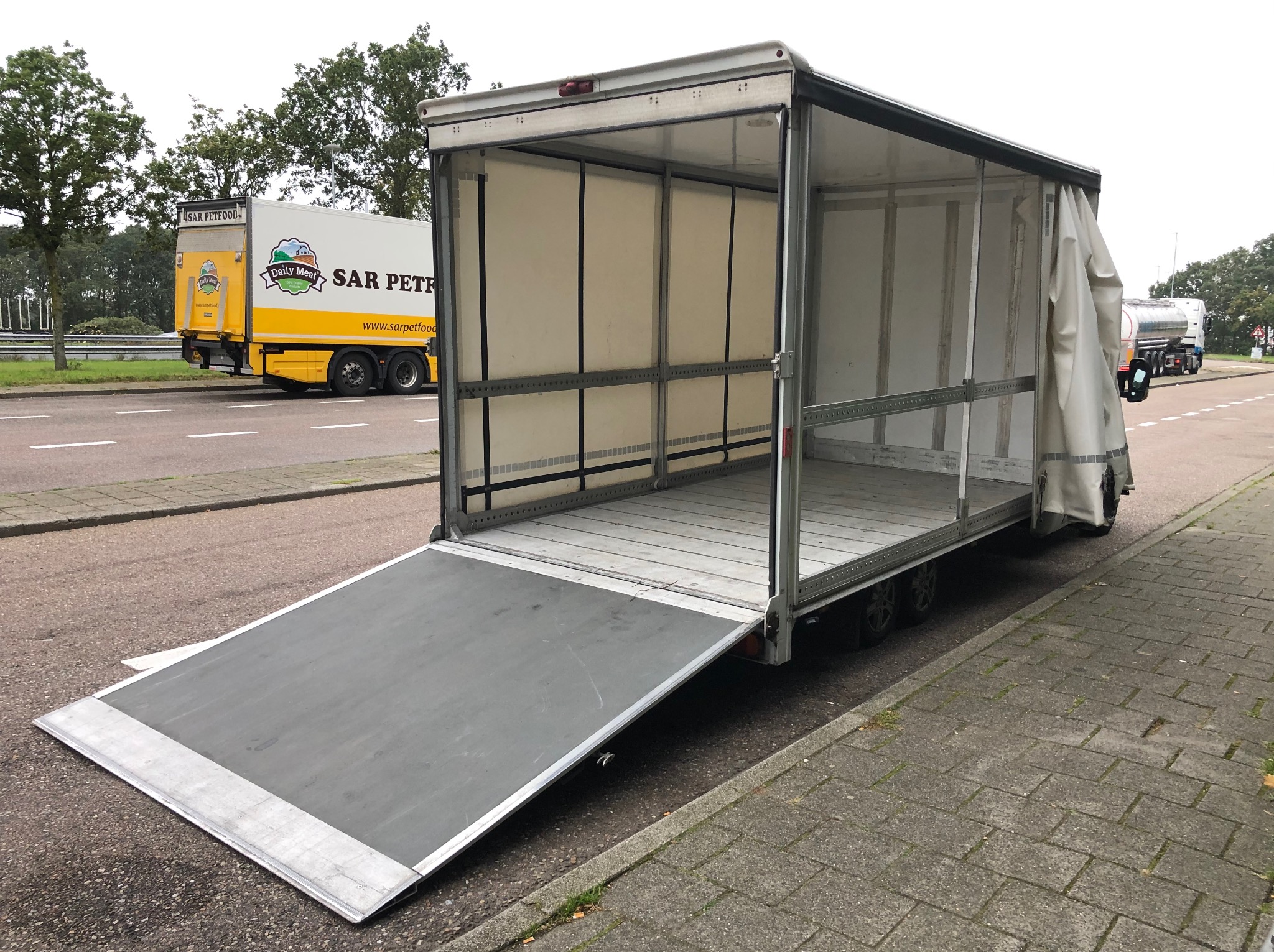 Otwarta paka busa z rampą na chodniku, widok z zewnątrz. W tle widać inne pojazdy na drodze. Rampa opuszczona, gotowa do załadunku.