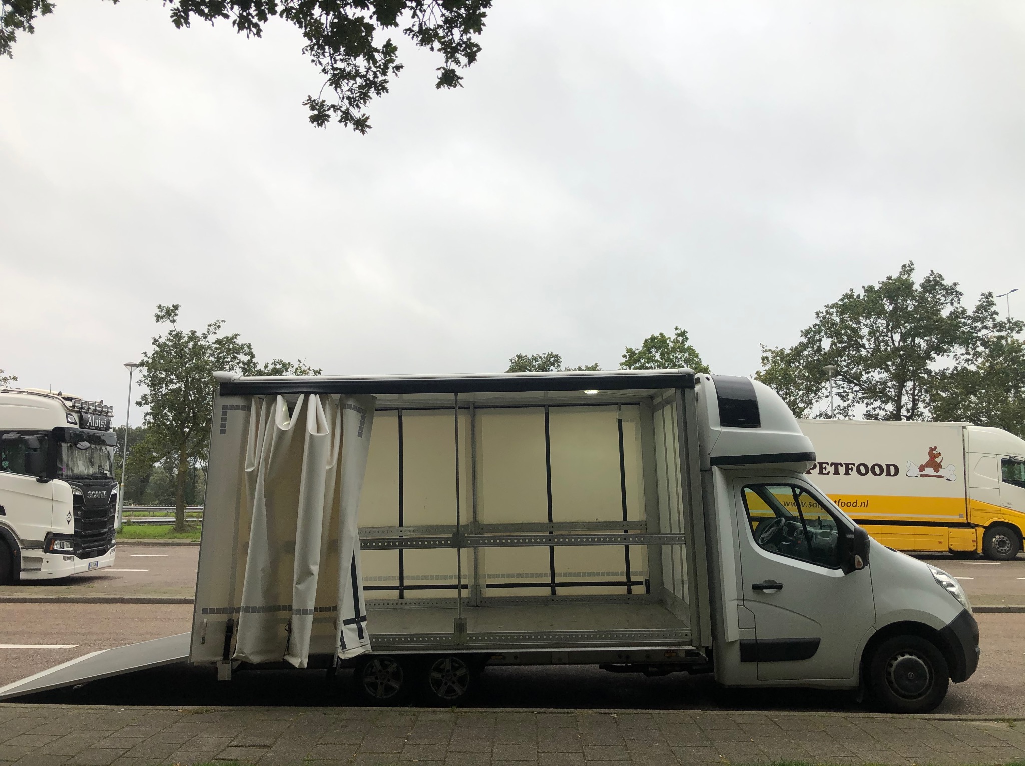Otwarty, pusty bus z rampą załadunkową i regałami wewnątrz, przygotowany do transportu. Obok zaparkowany tir z reklamą Petfood. Pochmurne niebo w tle.