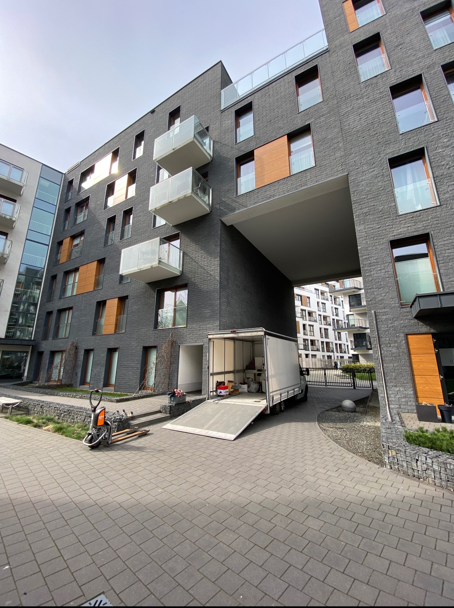 Przeprowadzka w nowoczesnym apartamentowcu z szarej cegły. Biała furgonetka z rampą i paleciak elektryczny na brukowanym podjeździe.