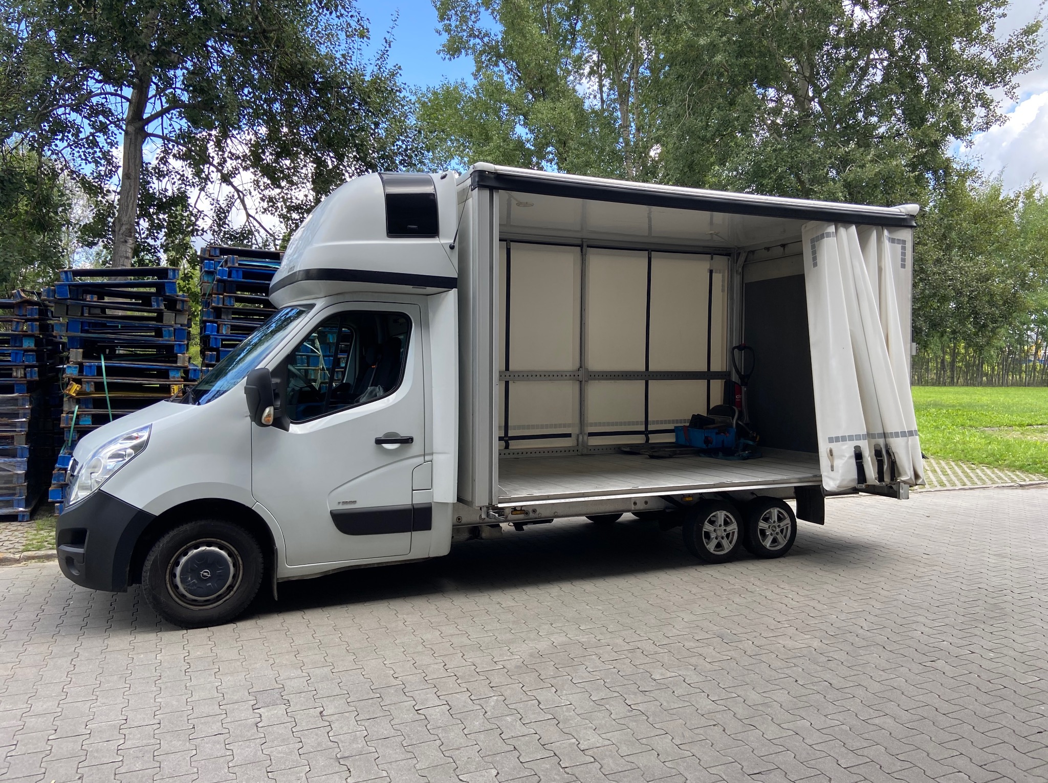 Biały bus z otwartą przestrzenią ładunkową na zewnątrz. W tle palety i zieleń. Widoczny wózek transportowy wewnątrz pojazdu. Bus zaparkowany na kostce brukowej.