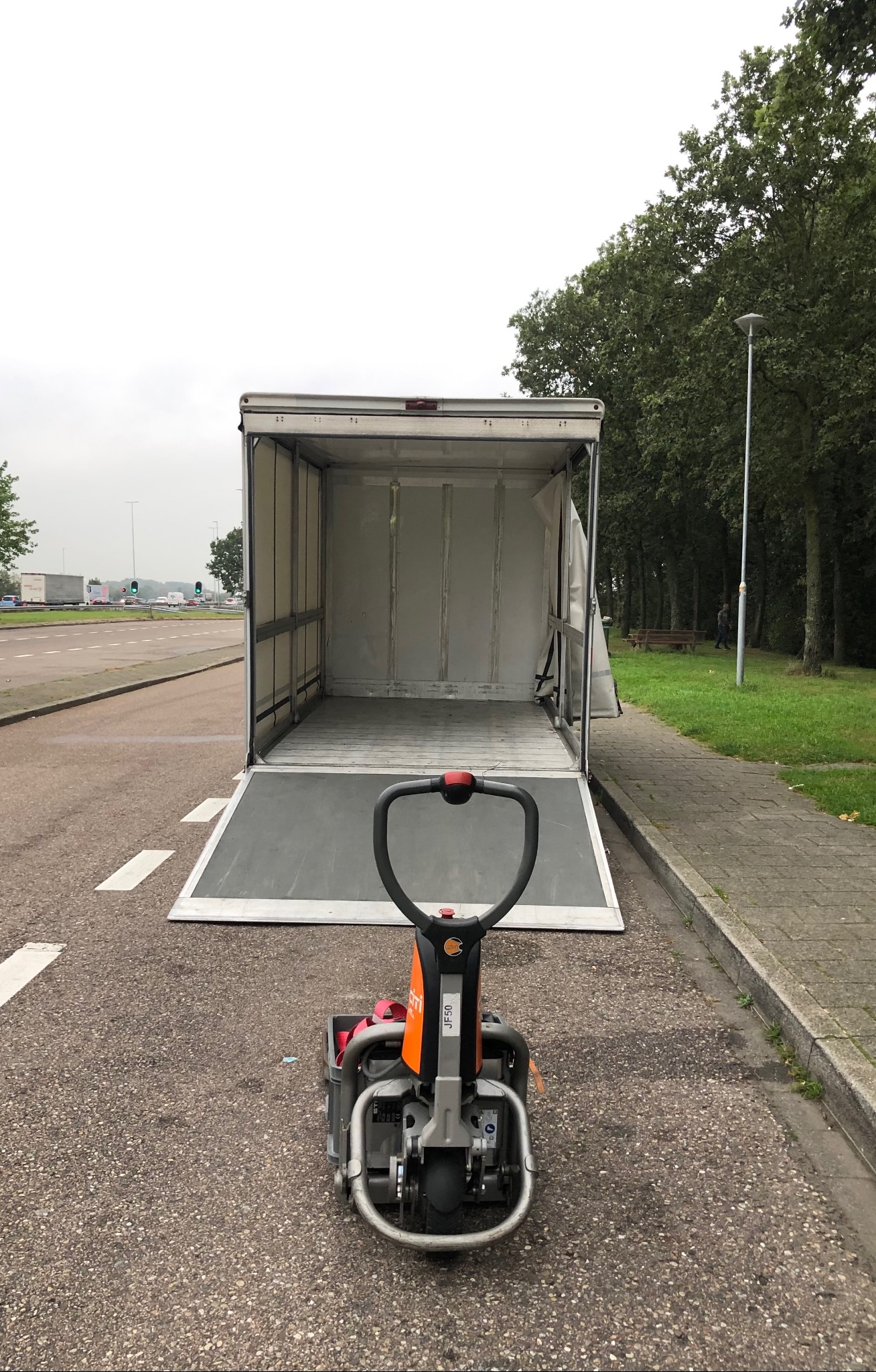 Otwarta paka busa z rampą załadowczą i elektrycznym wózkiem paletowym na asfalcie, w tle zieleń i droga. Sprzęt gotowy do załadunku towarów.