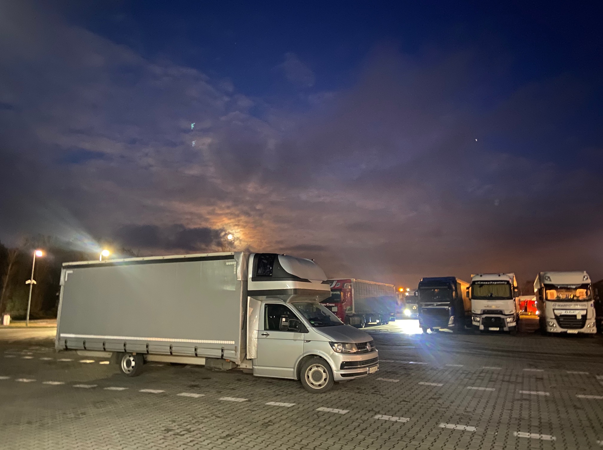 Srebrny bus dostawczy z zabudową kontenerową na parkingu, w tle inne samochody ciężarowe i nocne niebo z chmurami.