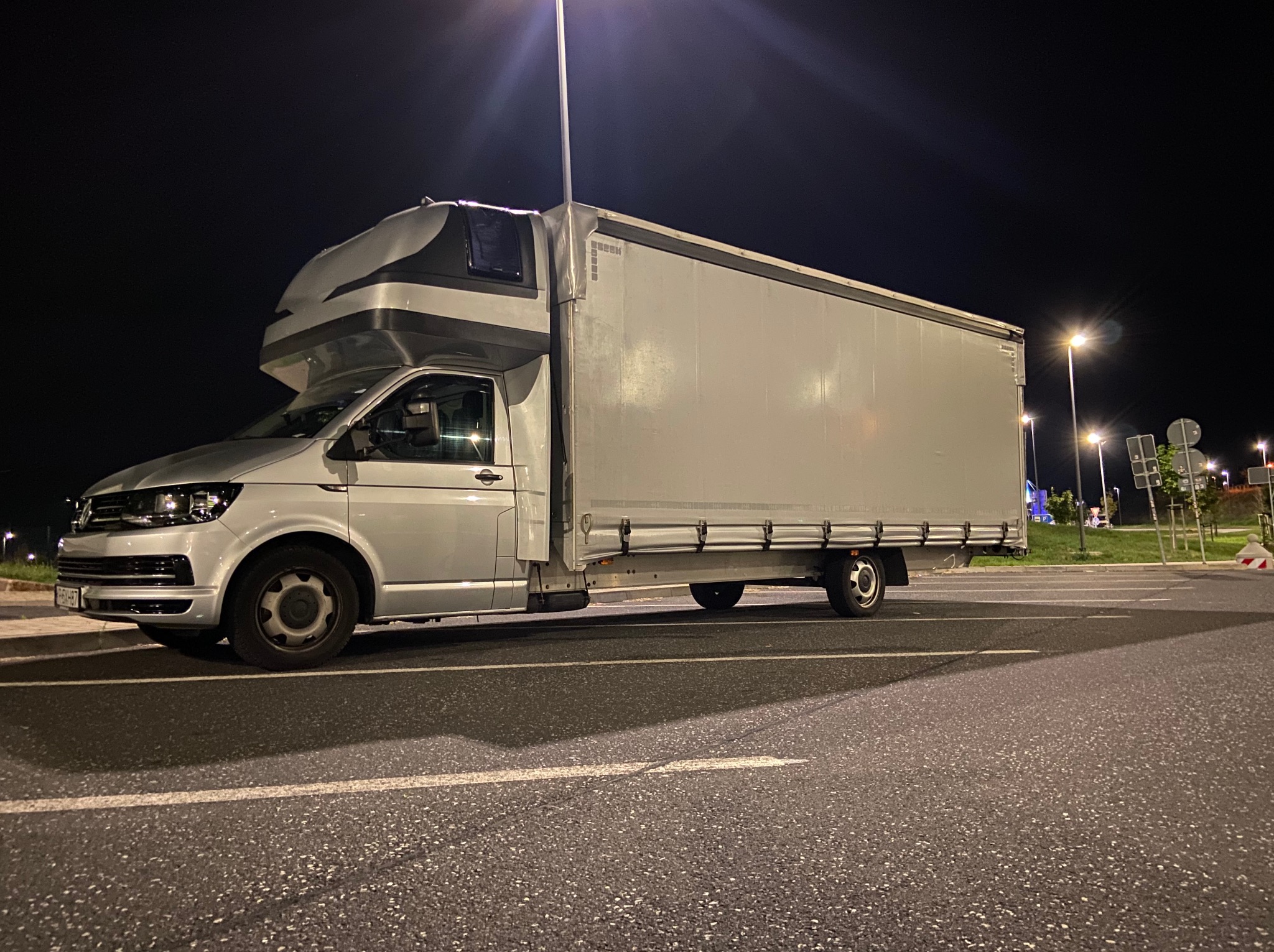 Srebrny bus z zabudową kontenerową parkuje nocą na parkingu oświetlonym latarniami. Nietypowa nadbudowa kabiny kierowcy. Widok z boku, światła uliczne w tle.