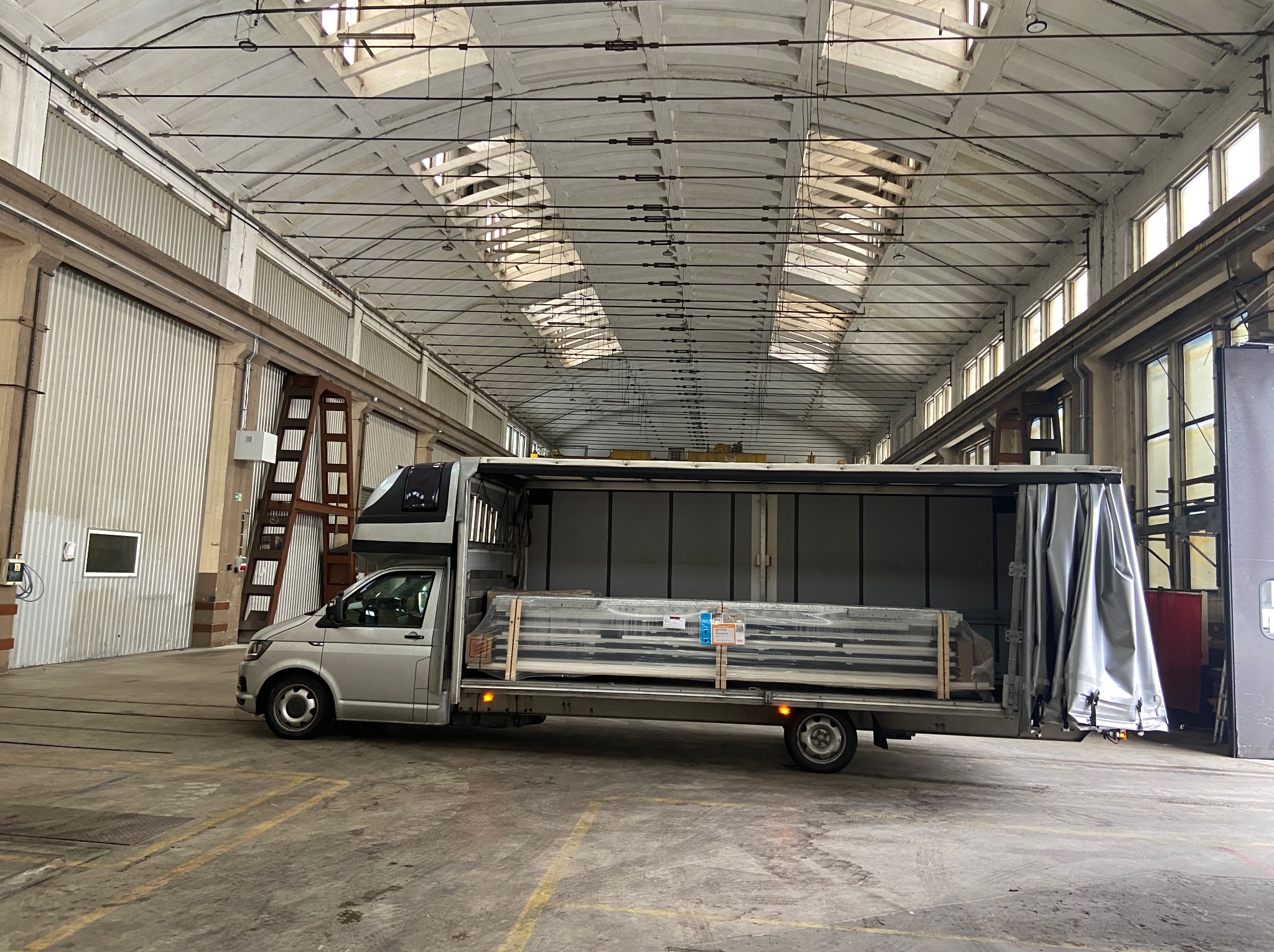 Srebrny bus z otwartą przestrzenią ładunkową, wypełnioną paletami z towarem, w hali magazynowej w Krakowie. Widoczne świetliki w suficie i betonowa podłoga.