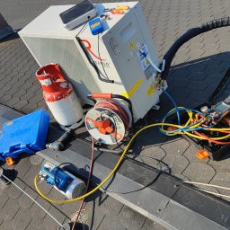 Jan Instalacje - Biała pompa ciepła na dachu z narzędziami: butla z gazem, wiertarka, zwinięty kabel, manometry. Widok z góry, dach pokryty dachówką bitumiczną, w tle zieleń i budynki.