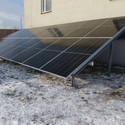 Jan Instalacje - Panel słoneczny zamontowany na śniegu przed budynkiem z oknem. Widoczne metalowe podpory i brązowe ogrodzenie w tle. Zimowy krajobraz.