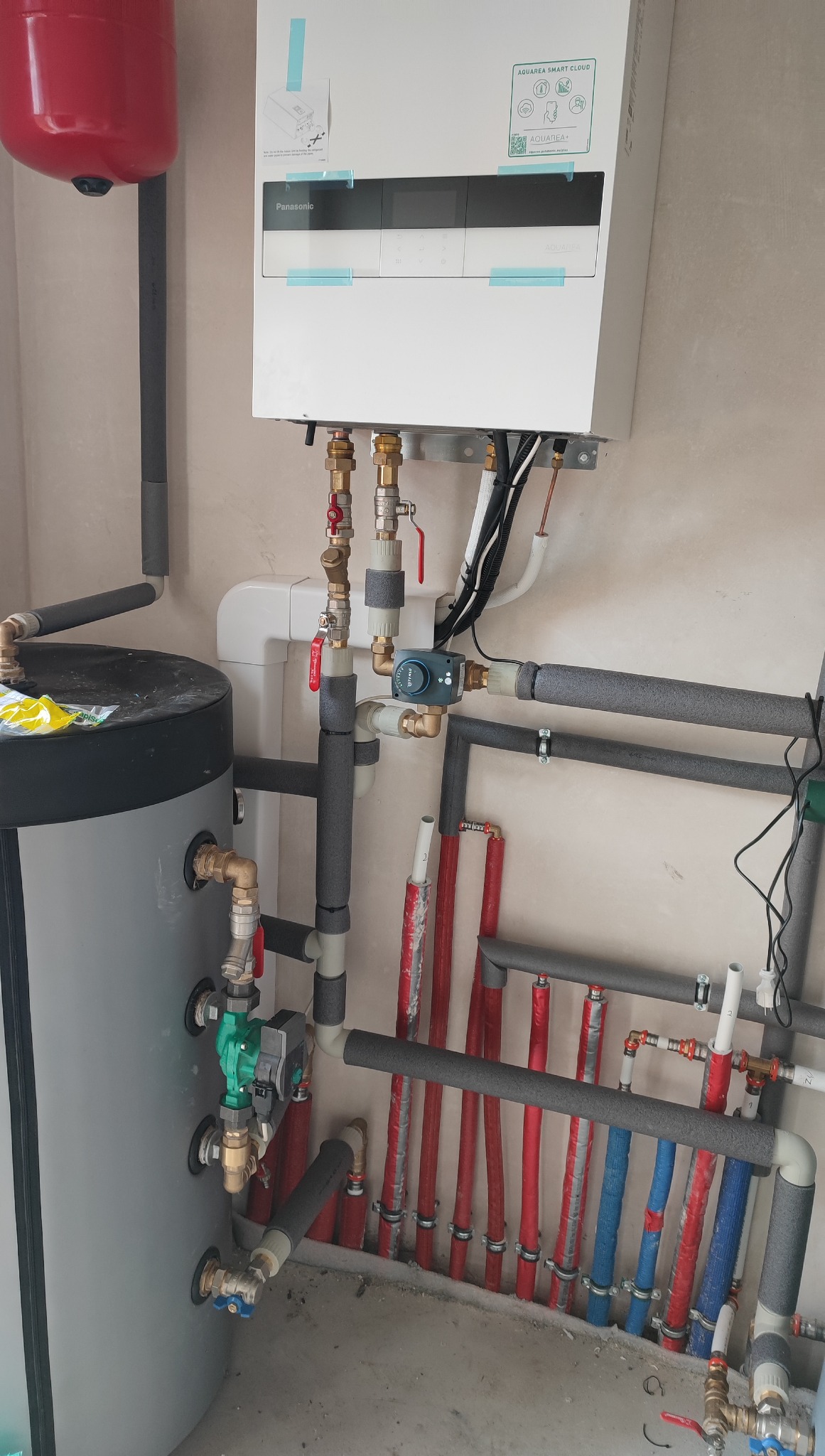 Złożona instalacja grzewcza z zbiornikiem, rurami w izolacji i panelem sterowania Panasonic Aquarea Smart Cloud, widoczna w pomieszczeniu technicznym.