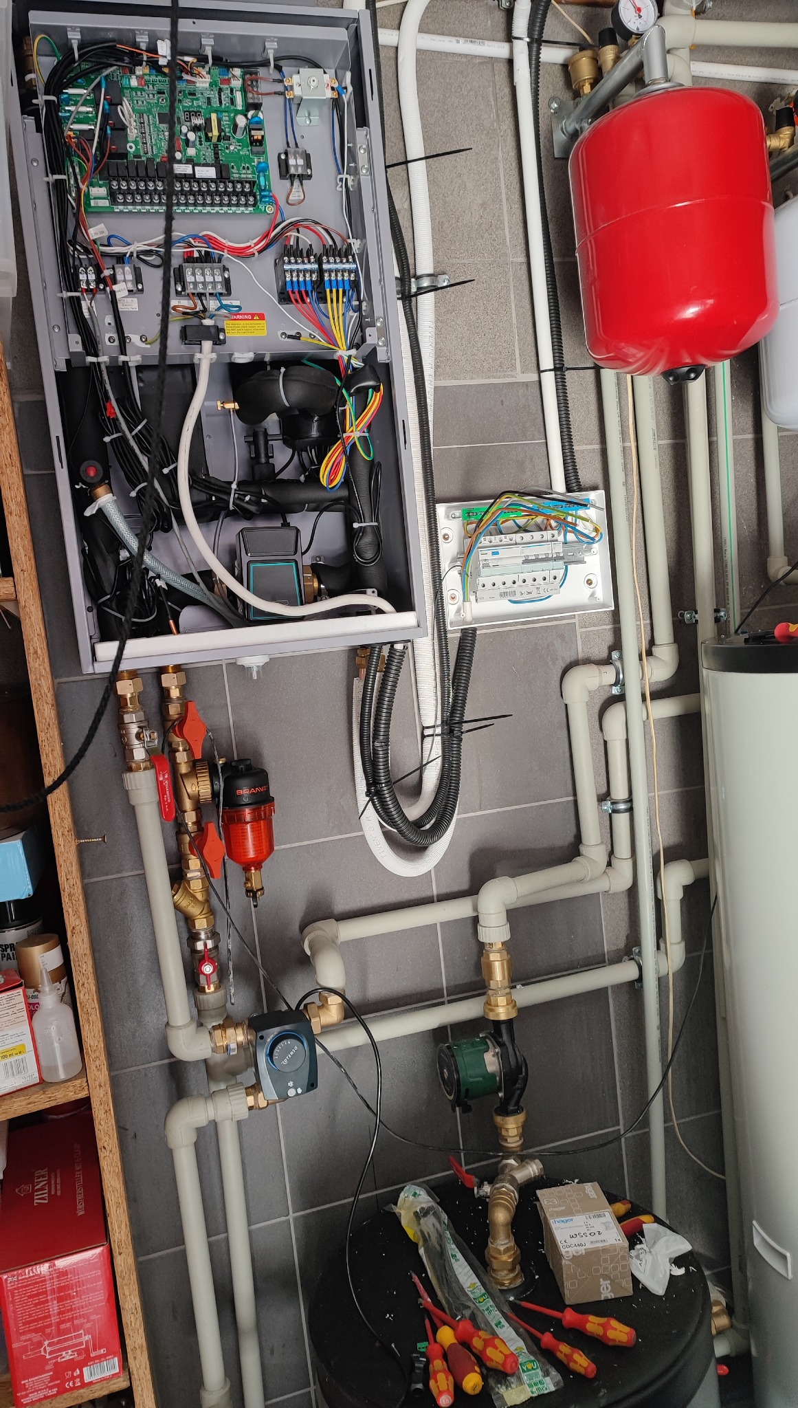 Rozbudowana instalacja hydrauliczna z licznymi rurami, zaworami, zbiornikiem wyrównawczym i otwartą skrzynką sterowniczą z elektroniką na szarej, kafelkowanej ścianie.