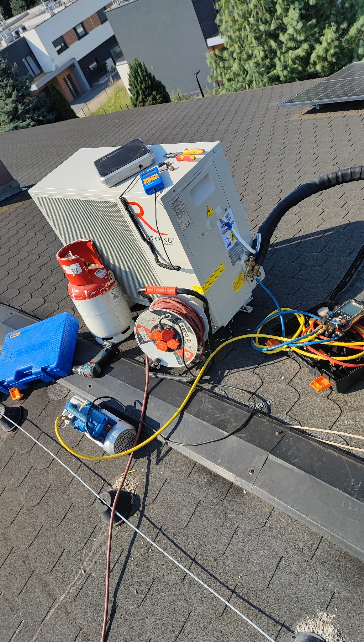 Biała pompa ciepła na dachu z narzędziami: butla z gazem, wiertarka, zwinięty kabel, manometry. Widok z góry, dach pokryty dachówką bitumiczną, w tle zieleń i budynki.