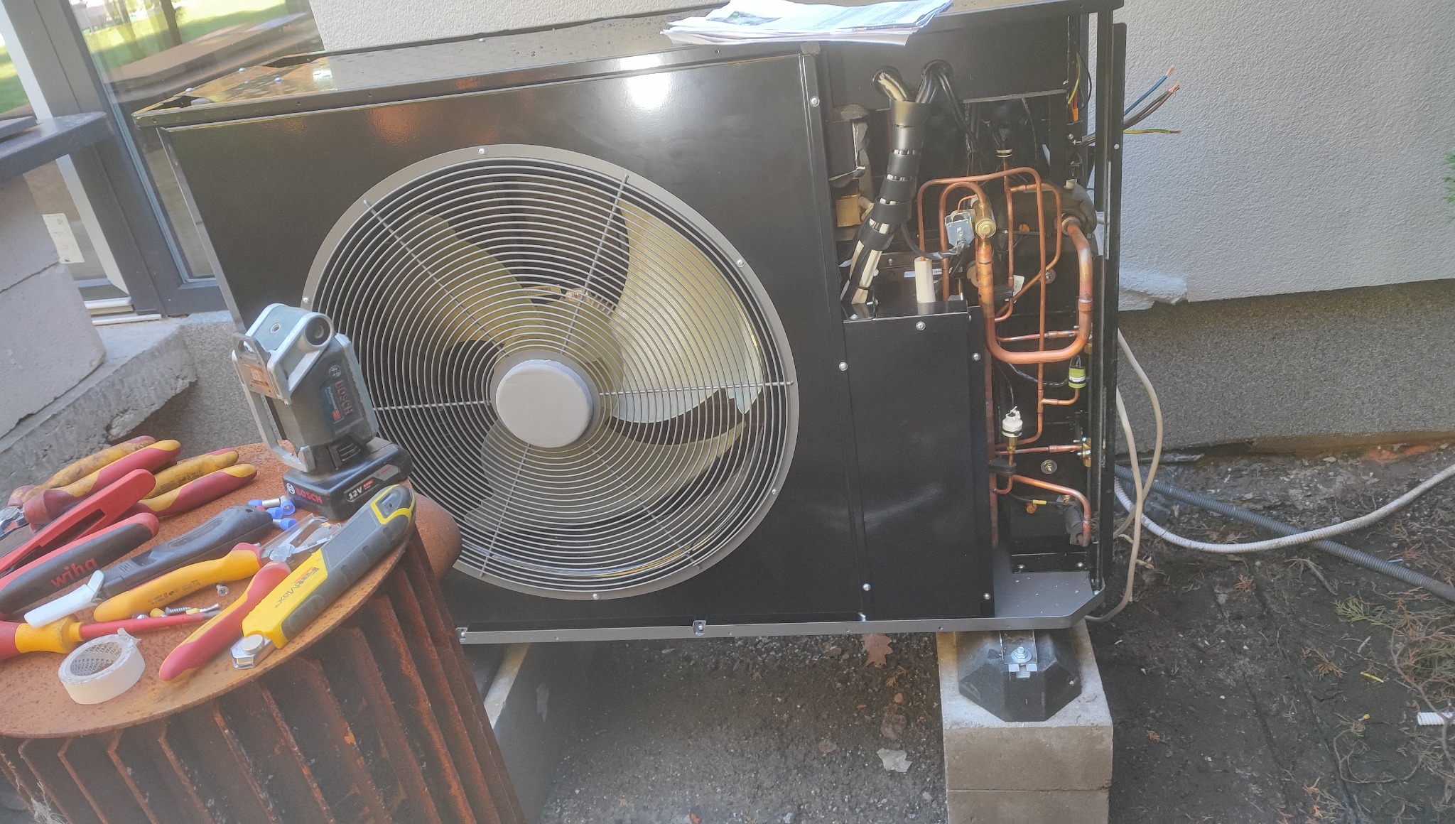 Otwarta obudowa pompy ciepła z widocznymi miedzianymi rurami i wentylatorem, narzędzia obok. Widoczne elementy instalacji i podłączenia elektryczne.