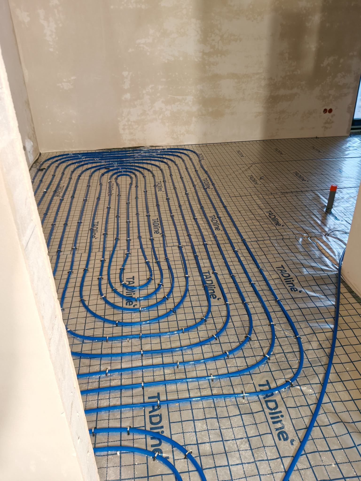 Niebieskie rury ogrzewania podłogowego ułożone na siatce w surowym wnętrzu, gotowe do zalania posadzką. Widoczne detale mocowań i oznaczenia producenta rur.