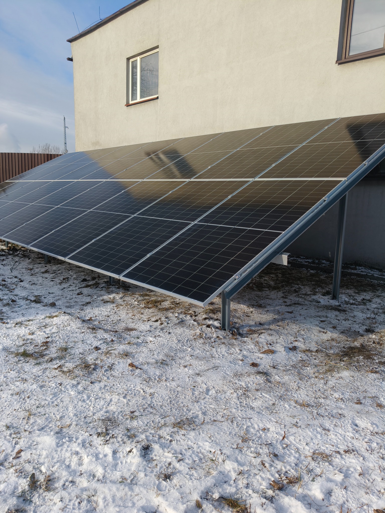 Panel słoneczny zamontowany na śniegu przed budynkiem z oknem. Widoczne metalowe podpory i brązowe ogrodzenie w tle. Zimowy krajobraz.