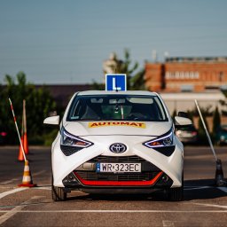 Turbo Szkolenia Michał Bogatek - Biały samochód z oznaczeniem 'L' i napisem 'Automat' na placu manewrowym szkoły jazdy, widok z przodu, z pomarańczowymi pachołkami i tyczkami po bokach.