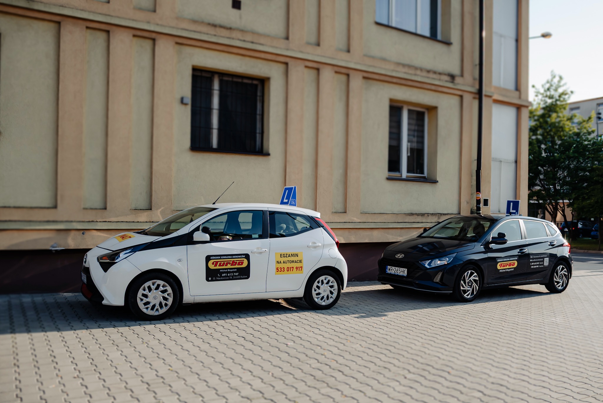 Dwa auta szkoły jazdy Turbo: białe Toyota Aygo z napisem 'Egzamin na automacie' i czarne Hyundai i20, zaparkowane przed budynkiem. Nauka jazdy.