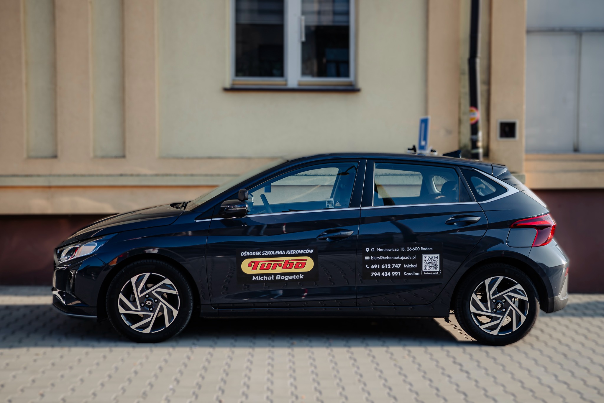 Czarny samochód z logo 'Turbo' szkoły jazdy, zaparkowany przed budynkiem. Widoczny adres i dane kontaktowe szkoły na boku pojazdu.
