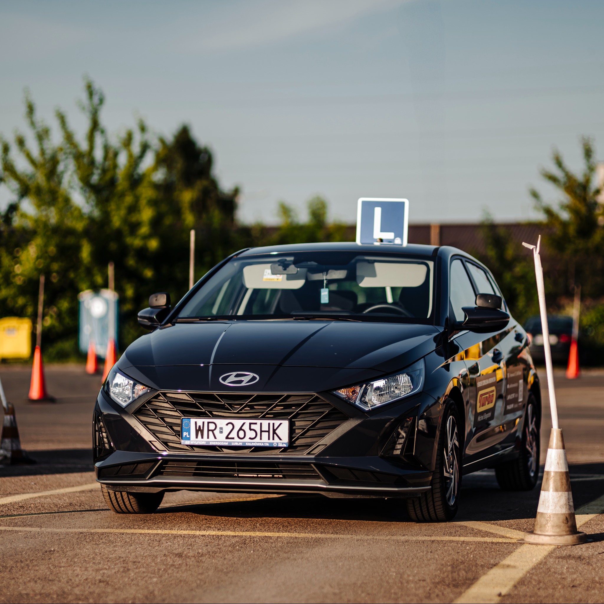 Czarny samochód nauki jazdy z literą 'L' na dachu, zaparkowany na placu manewrowym z pachołkami i liniami. Widoczne logo i tablica rejestracyjna.