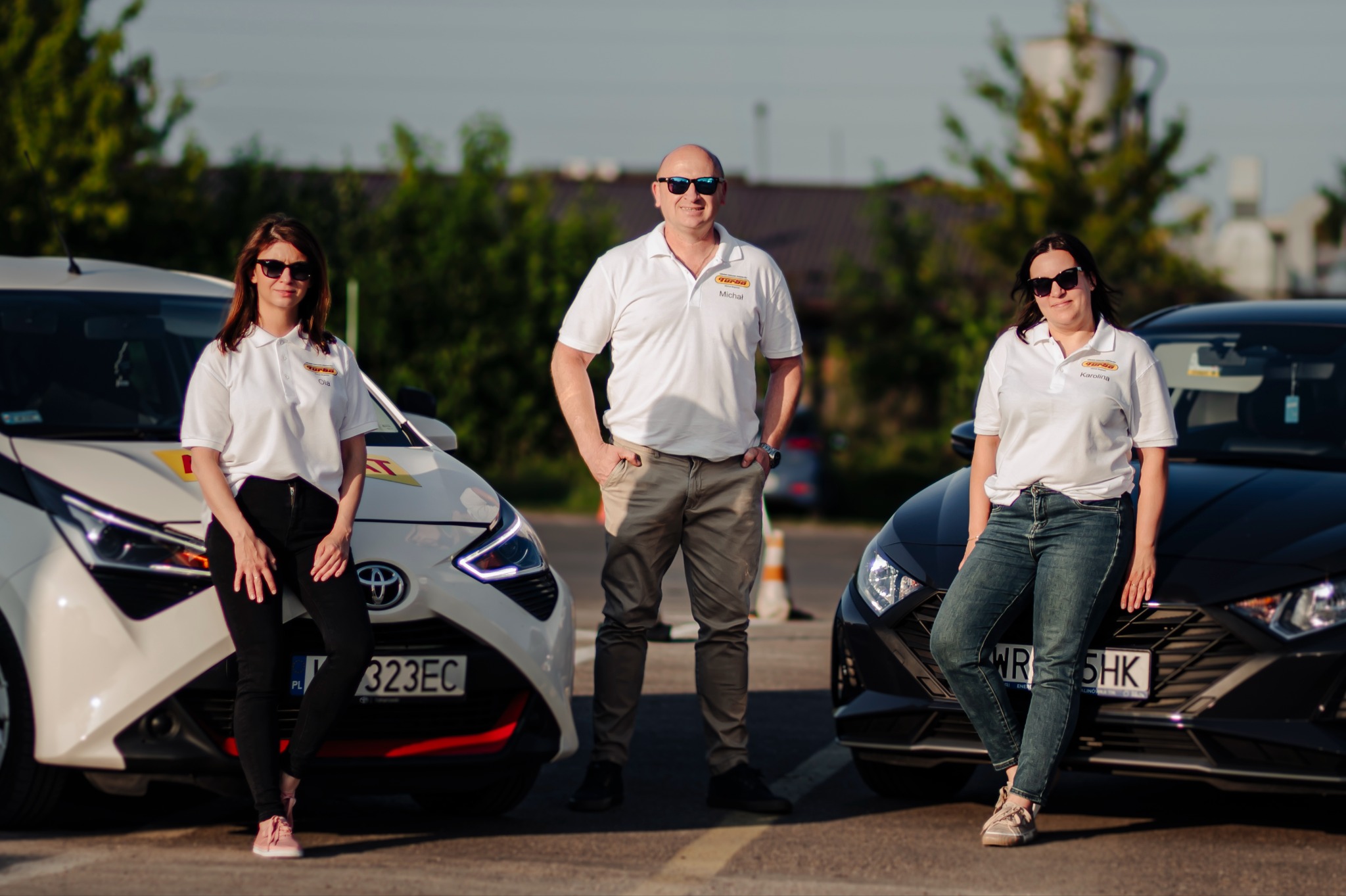 Instruktorzy szkoły jazdy w koszulkach polo z logo, stojący przed samochodami nauki jazdy. Dwie kobiety opierają się o maski aut, mężczyzna w środku pozuje z rękami w kieszeniach.