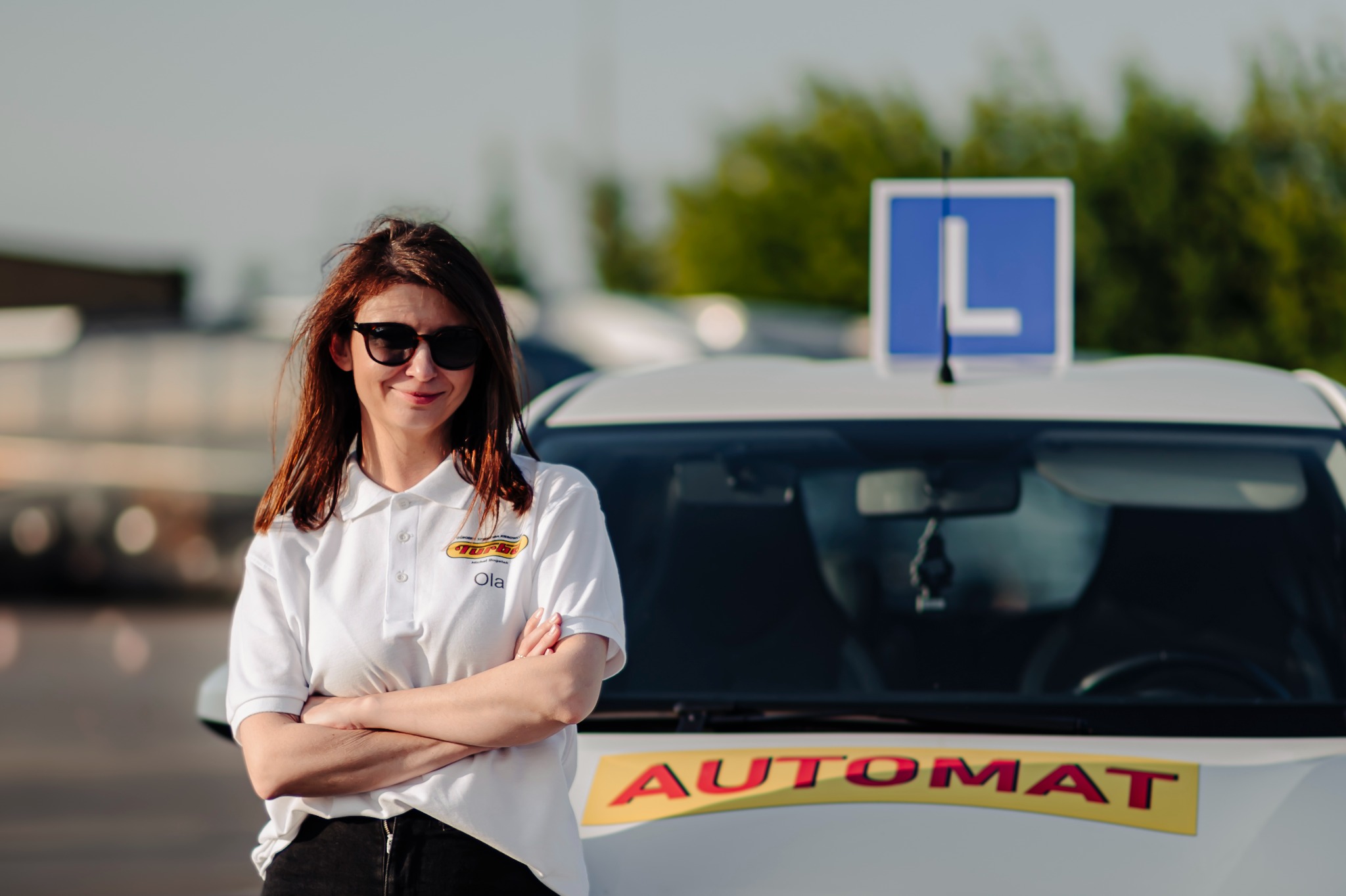 Instruktorka 'Ola' w okularach, oparta o auto z napisem 'Automat' i 'L' na dachu. Uśmiechnięta, skrzyżowane ręce, biała koszulka polo z logo szkoły jazdy.