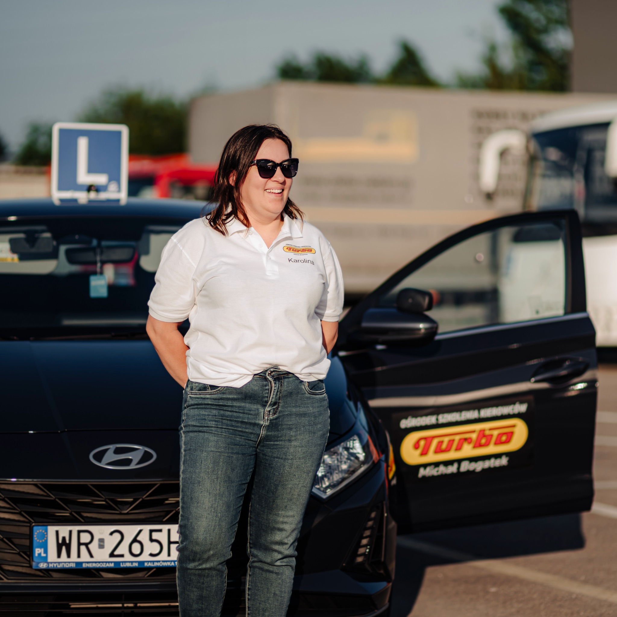 Instruktorka 'Turbo' uśmiecha się obok auta z otwartymi drzwiami i znakiem 'L'. Tablica rejestracyjna pojazdu WR 265H. Widoczny budynek w tle.