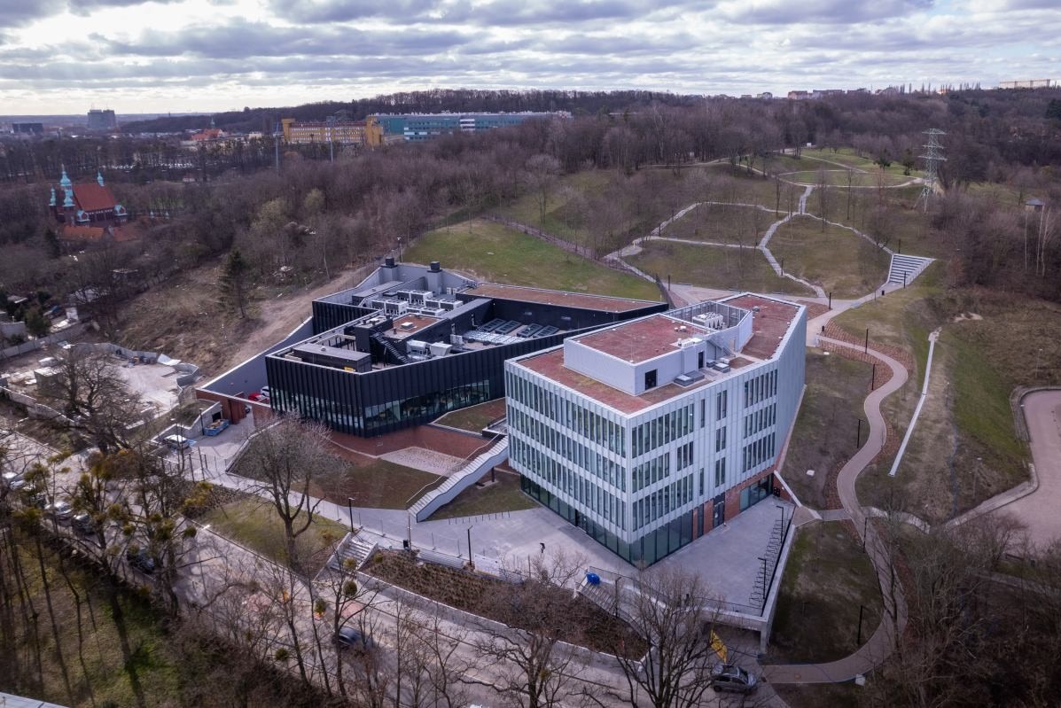 Zdjęcie z drona ukazuje nowoczesny kompleks budynków z zielonymi dachami, otoczony parkiem i ścieżkami spacerowymi. Widoczny kościół w tle. Pochmurne niebo.