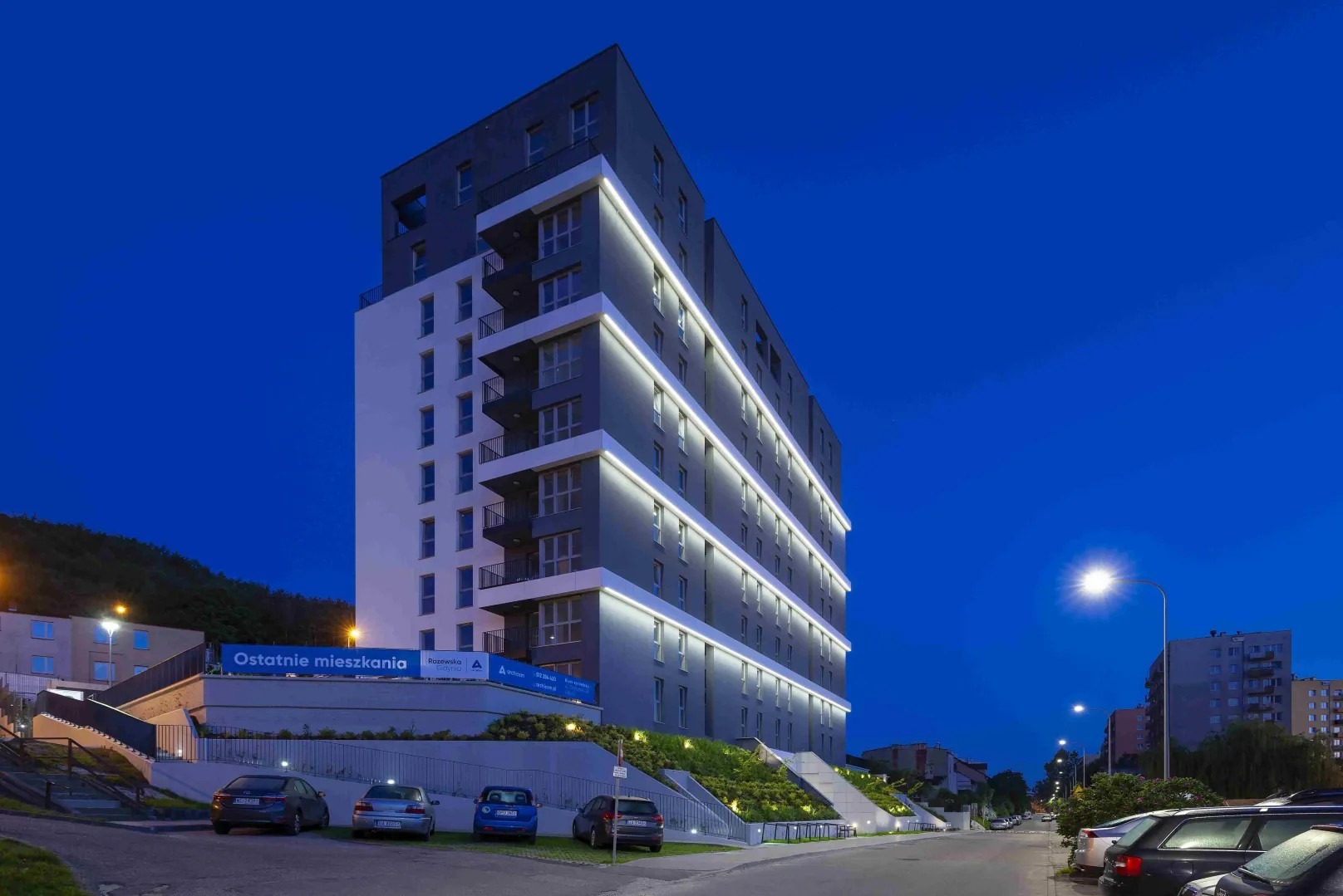 Wielorodzinny budynek mieszkalny z oświetleniem elewacji, widoczny z poziomu ulicy o zmierzchu. Uwagę zwraca nowoczesna architektura i podświetlenie balkonów.