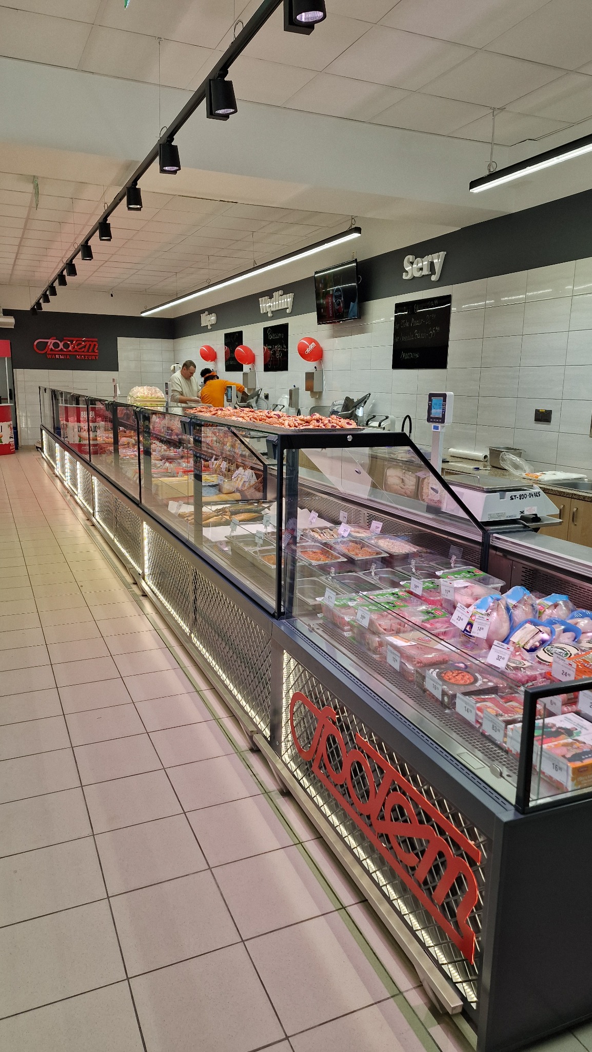 Długa, podświetlana witryna chłodnicza z różnorodnymi produktami mięsnymi i wędliniarskimi w sklepie. Widoczny personel i dekoracje balonowe.
