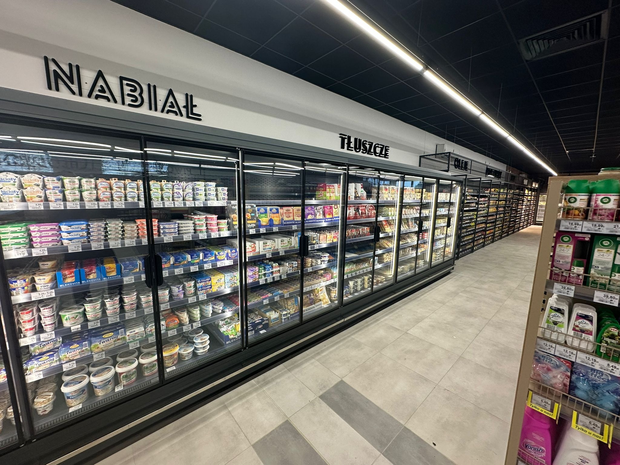 Długi rząd przeszklonych chłodziarek z nabiałem i tłuszczami w supermarkecie, oświetlony ledowymi lampami sufitowymi.