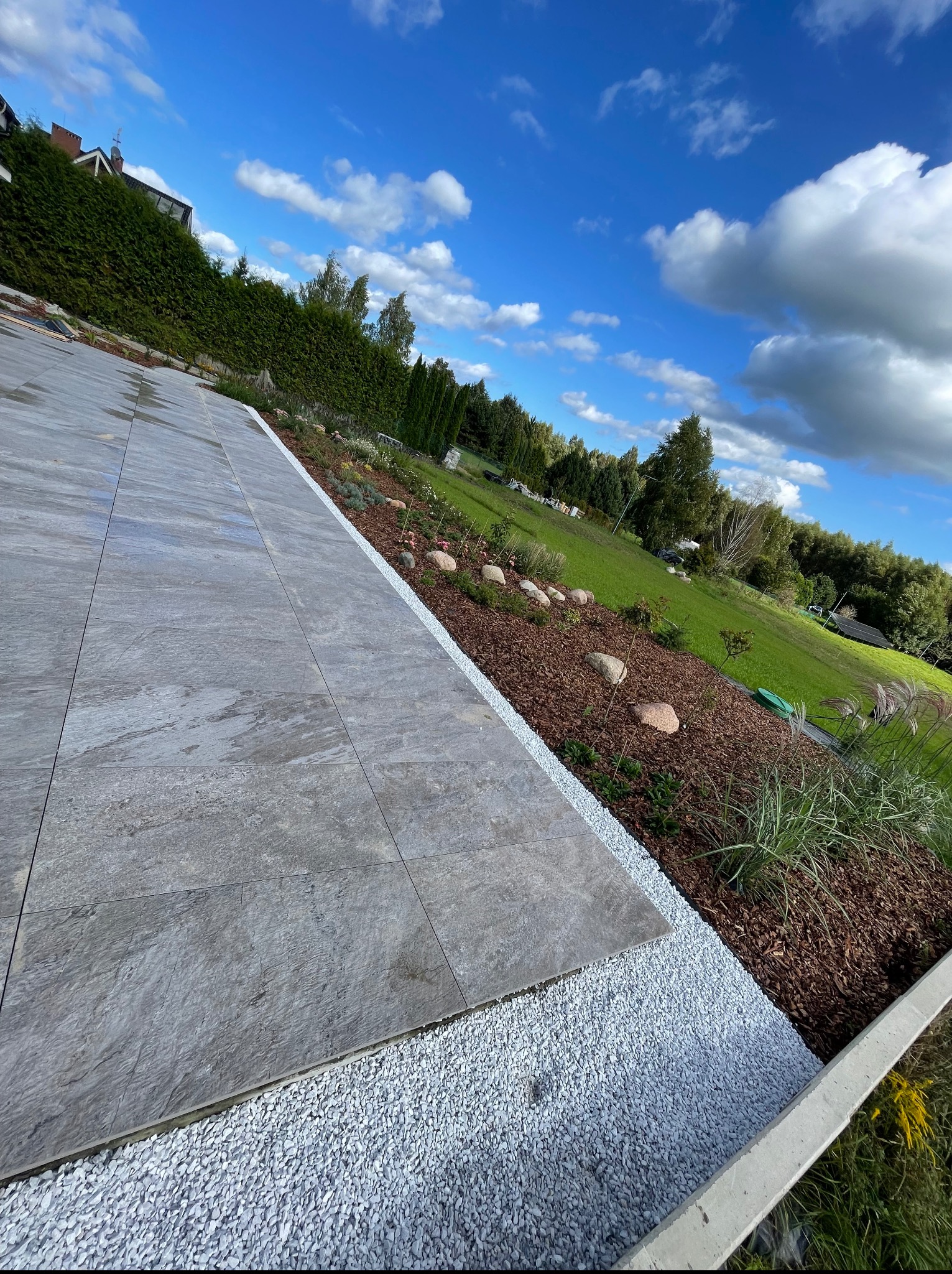 Nowoczesny ogród z szarym brukiem, białym żwirem i korą drzewną. Ujęcie ukośne, podkreślające perspektywę i głębię ogrodu w słoneczny dzień.