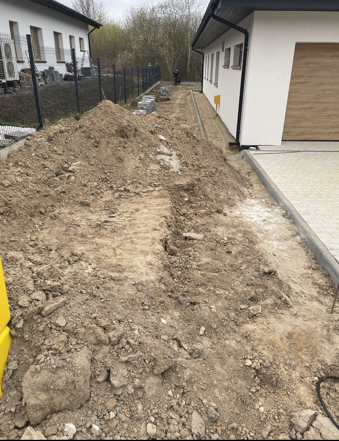 Przygotowanie terenu pod budowę chodnika z kostki brukowej. Widoczna wykopana ziemia, obrzeża i częściowo ułożona kostka. Prace w toku, dom w tle.