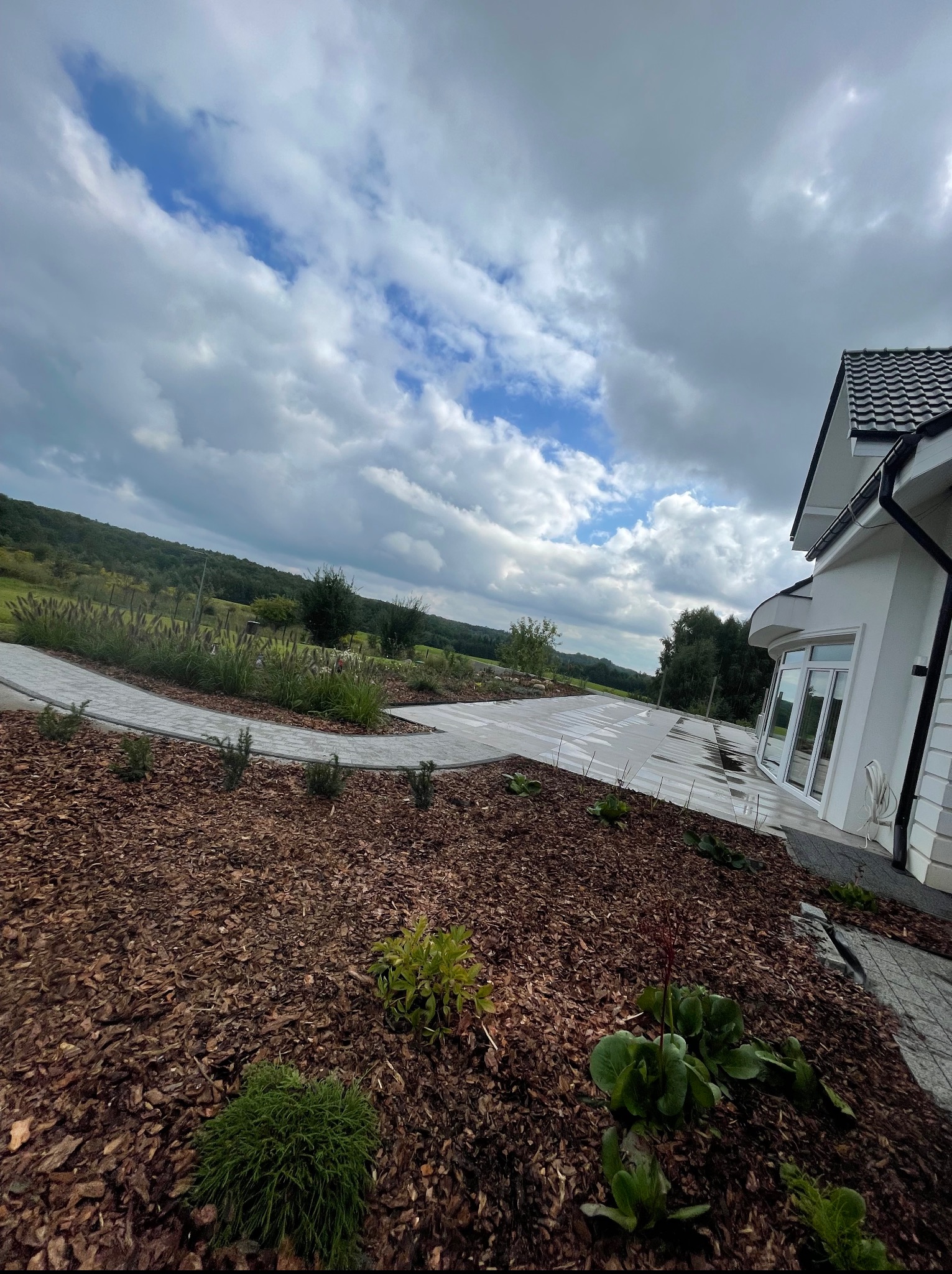 Nowoczesny ogród z ścieżką z kostki brukowej, rabatami wysypanymi korą i widokiem na malowniczy krajobraz. Biały dom z dużymi oknami w tle.