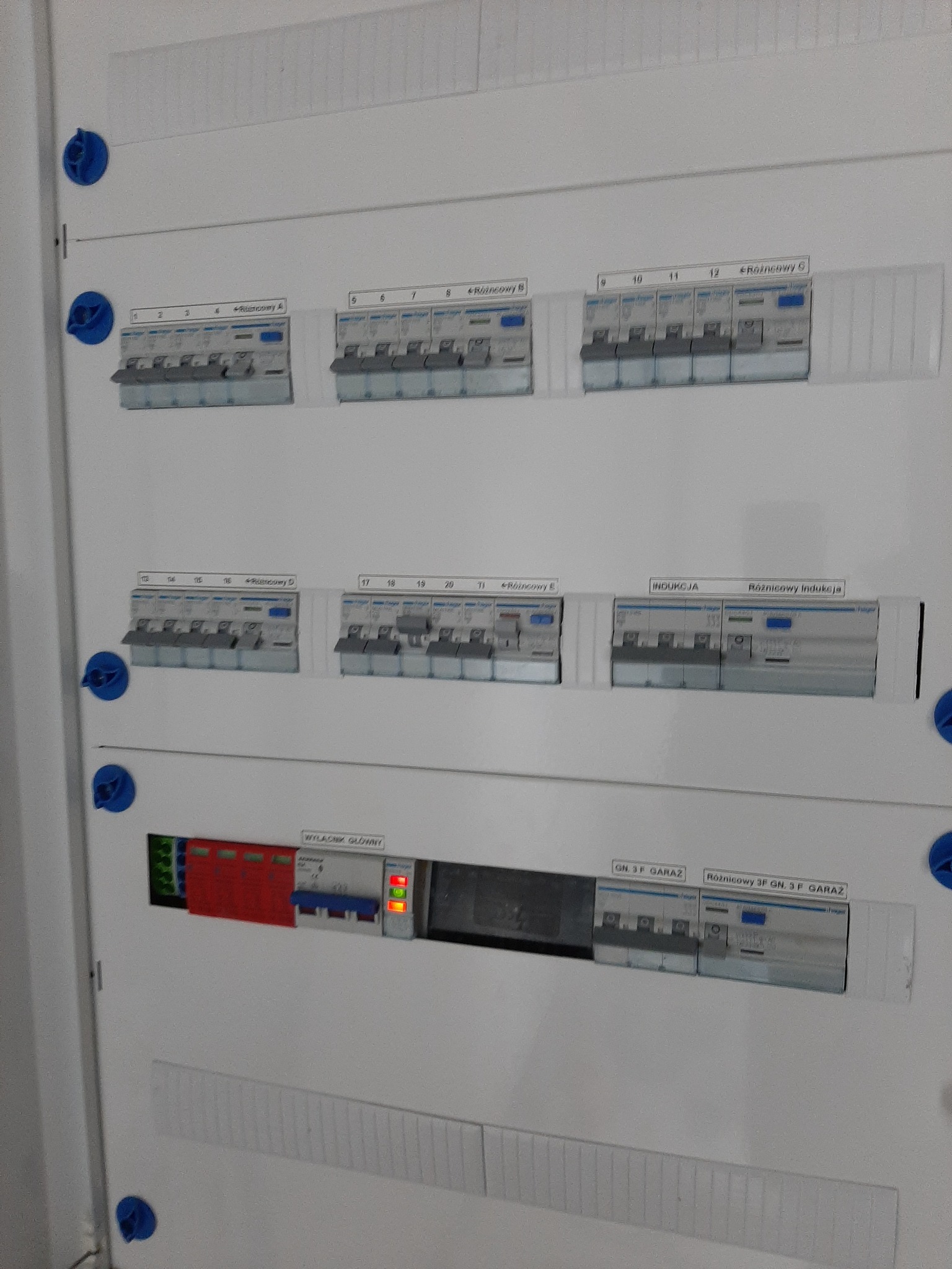Rozdzielnica elektryczna z bezpiecznikami różnicowoprądowymi i wyłącznikiem głównym. Widoczne opisy obwodów: 'Różnicowy A, B, C, D, E', 'Indukcja', 'Garaż'.