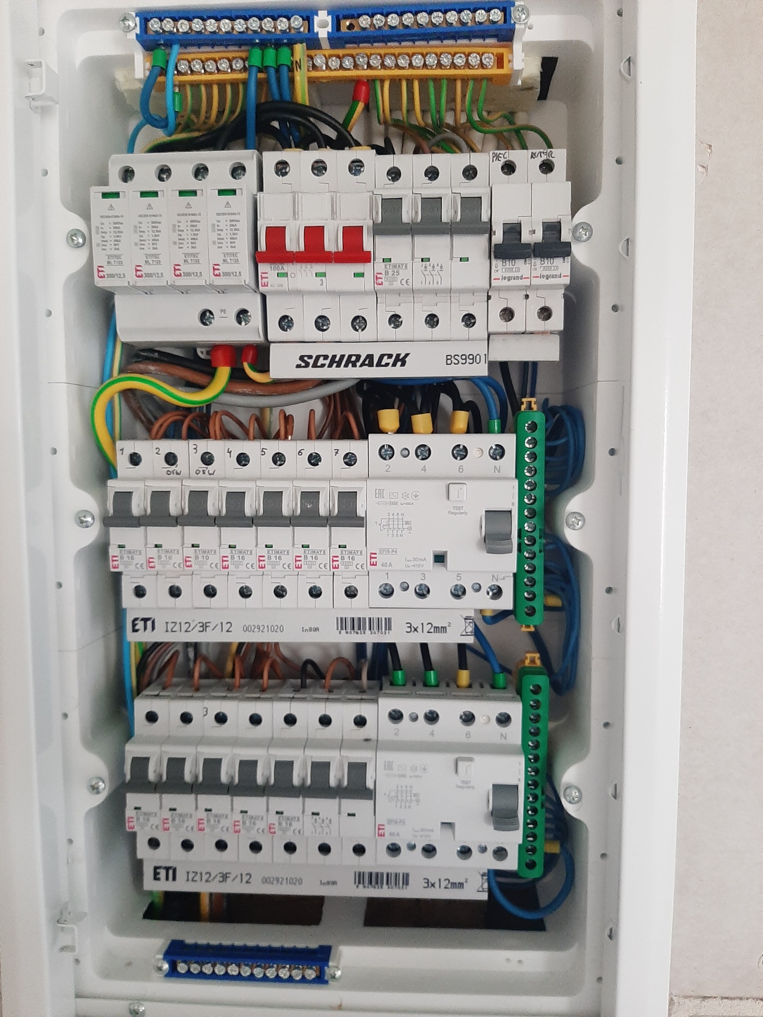 Rozdzielnica elektryczna w białej obudowie z widocznymi bezpiecznikami, przewodami i złączkami. Widoczne elementy marki Schrack i ETI.