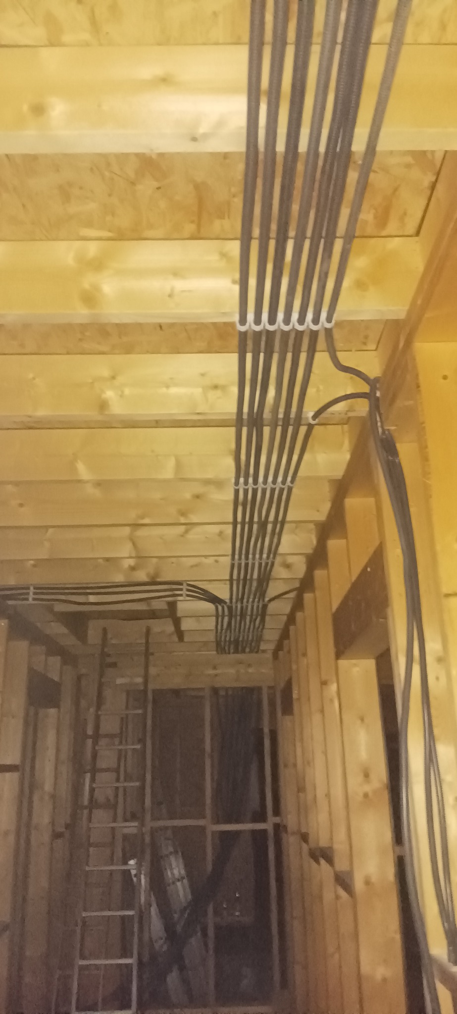 Układanie kabli w peszlach na drewnianym stropie podczas prac instalacyjnych w nowym budynku. Widoczna drabina i konstrukcja szkieletowa.